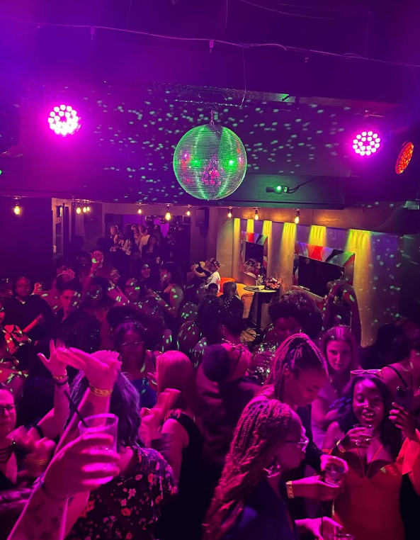 Crowd dancing in a club with purple and green lighting, a disco ball, and colorful wall lights.