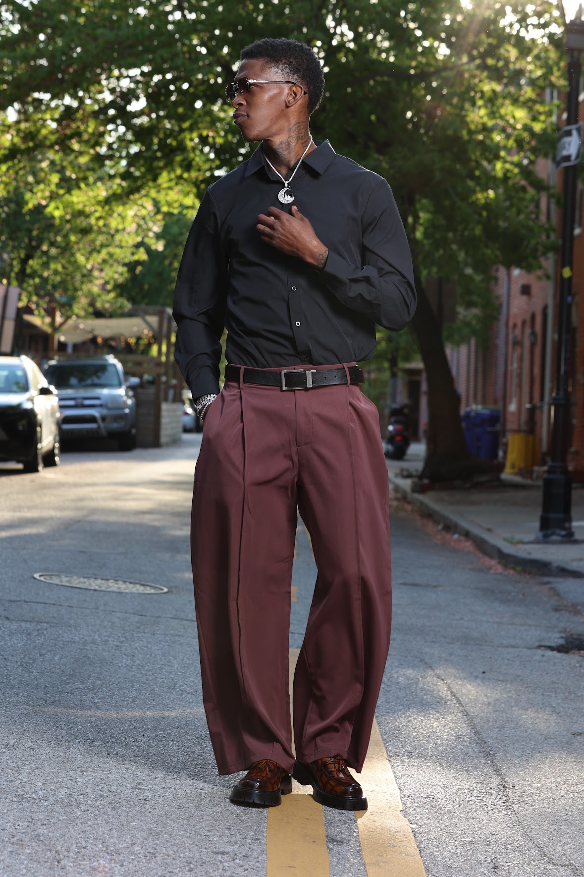 Person standing on street wearing black shirt, maroon pants, sunglasses, and headphones, with trees and cars in background.