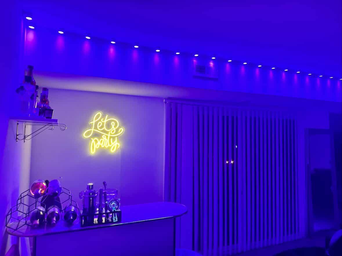 Room with blue lighting, neon sign reading 'let's party,' bar with bottles and glasses, and vertical blinds.