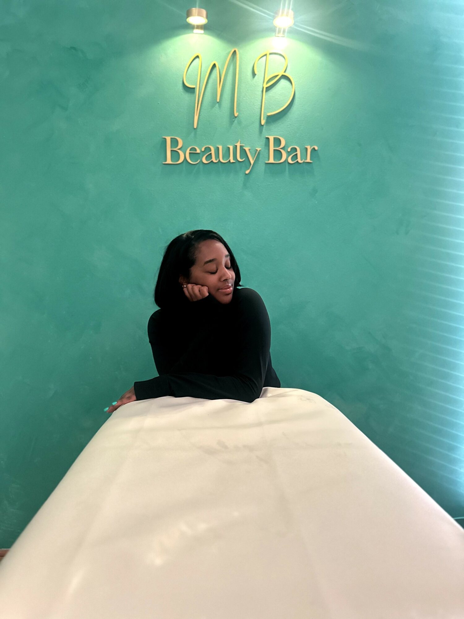 Woman sitting at a beauty bar with a green wall and gold sign reading 'MB Beauty Bar'.