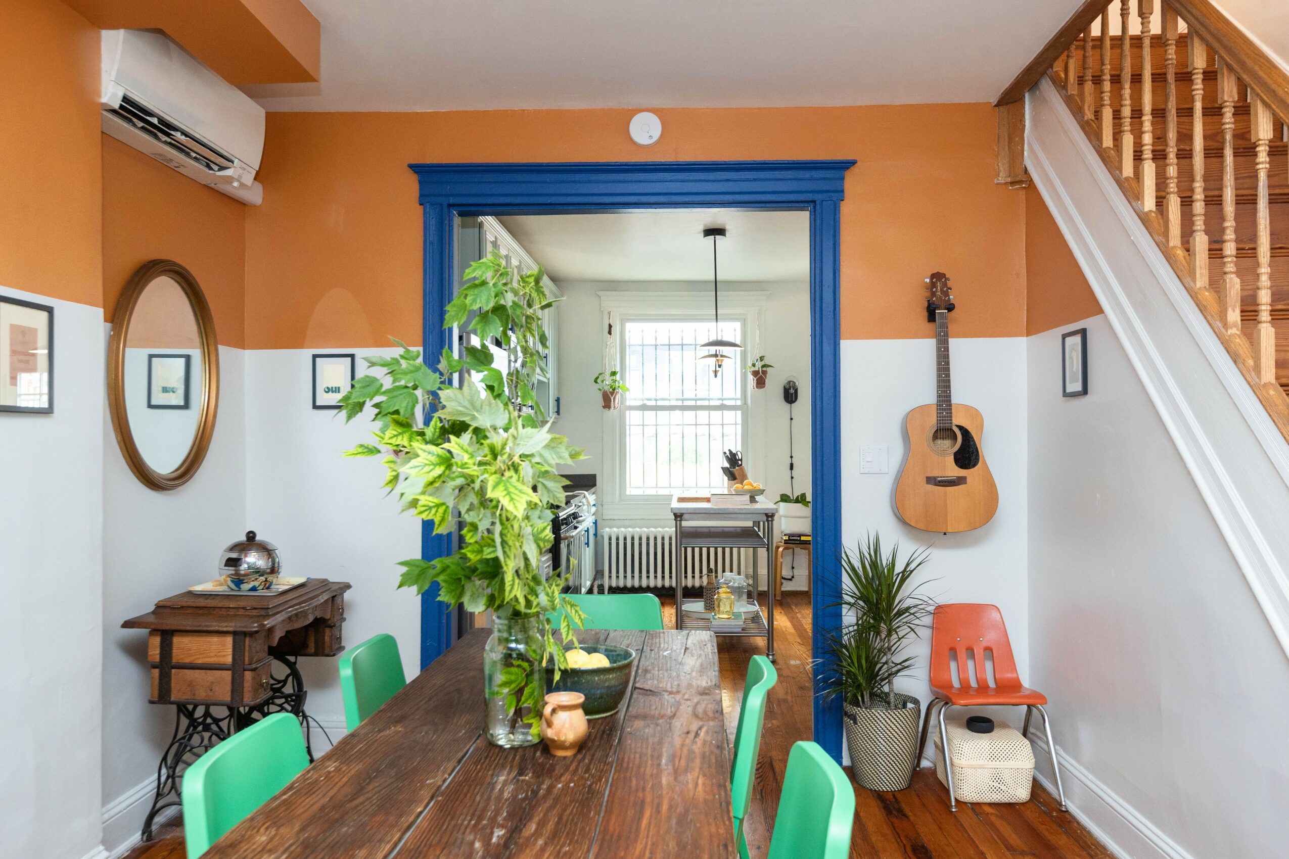 Dining room with a large plant on the table, blue door frame, guitar on wall, and staircase on right side.