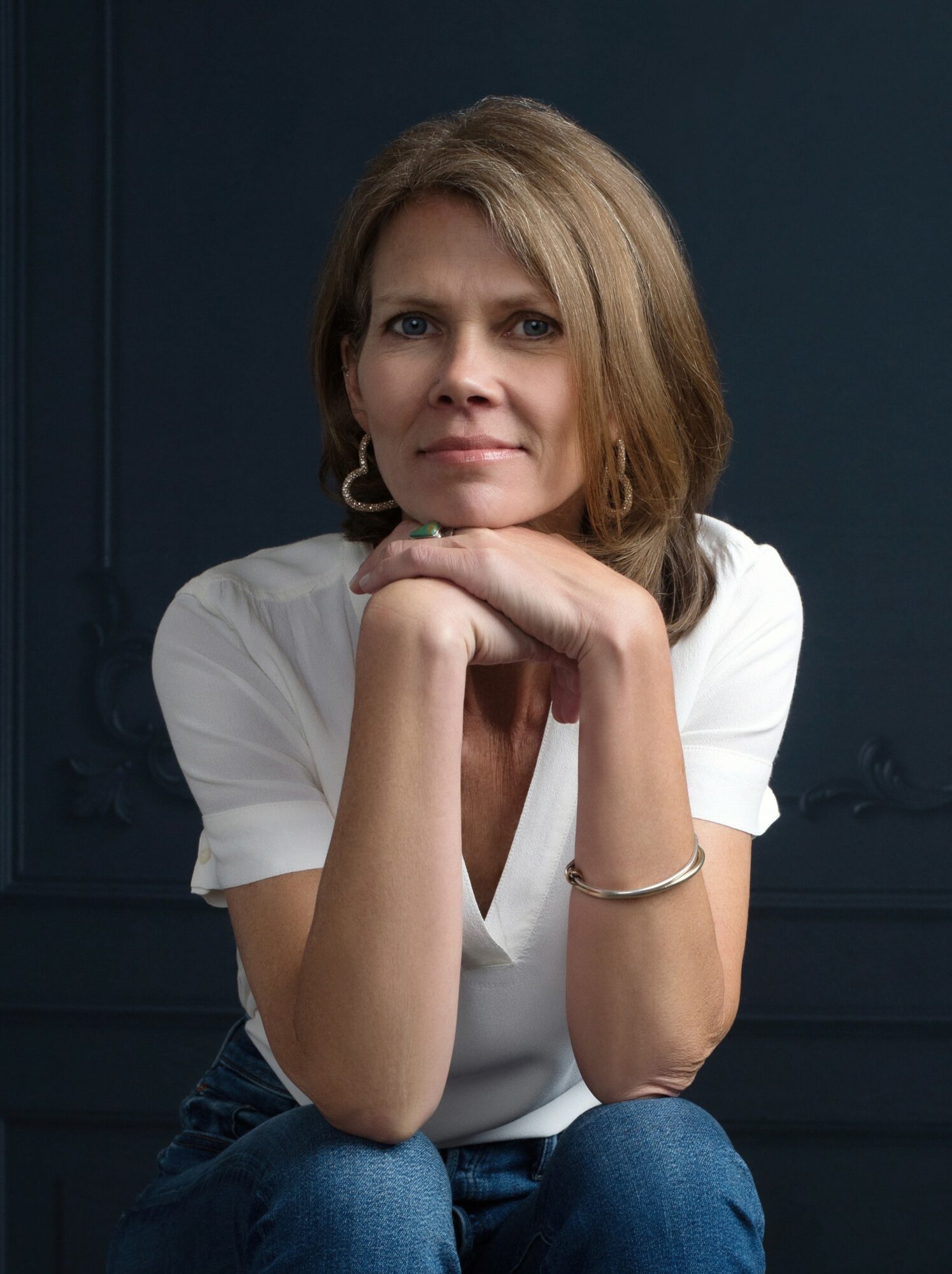 Woman with shoulder-length hair, wearing a white top, sitting with hands under chin, against dark background.