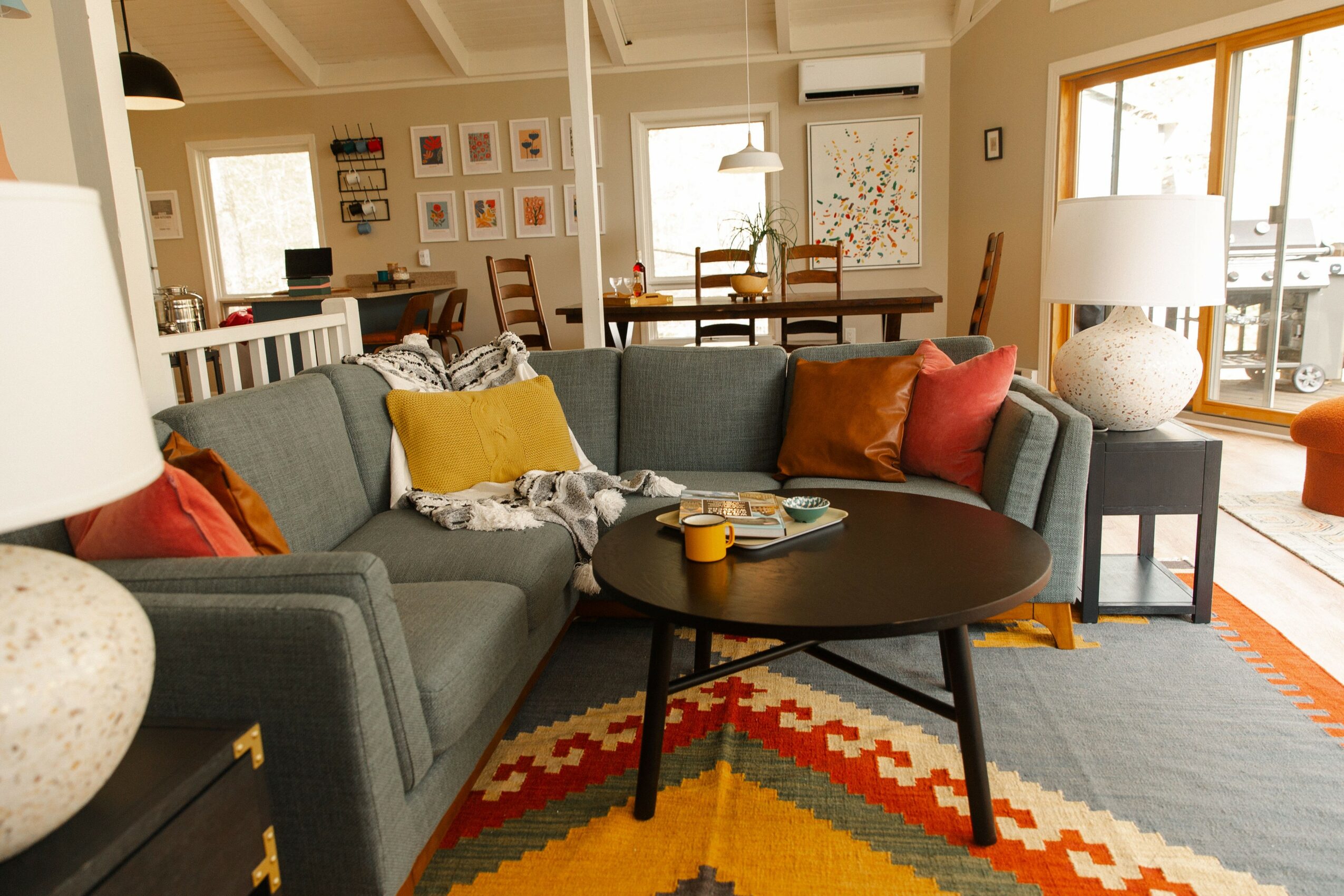 Living room with gray sectional sofa, colorful pillows, round coffee table, and a patterned rug, with dining area in background.