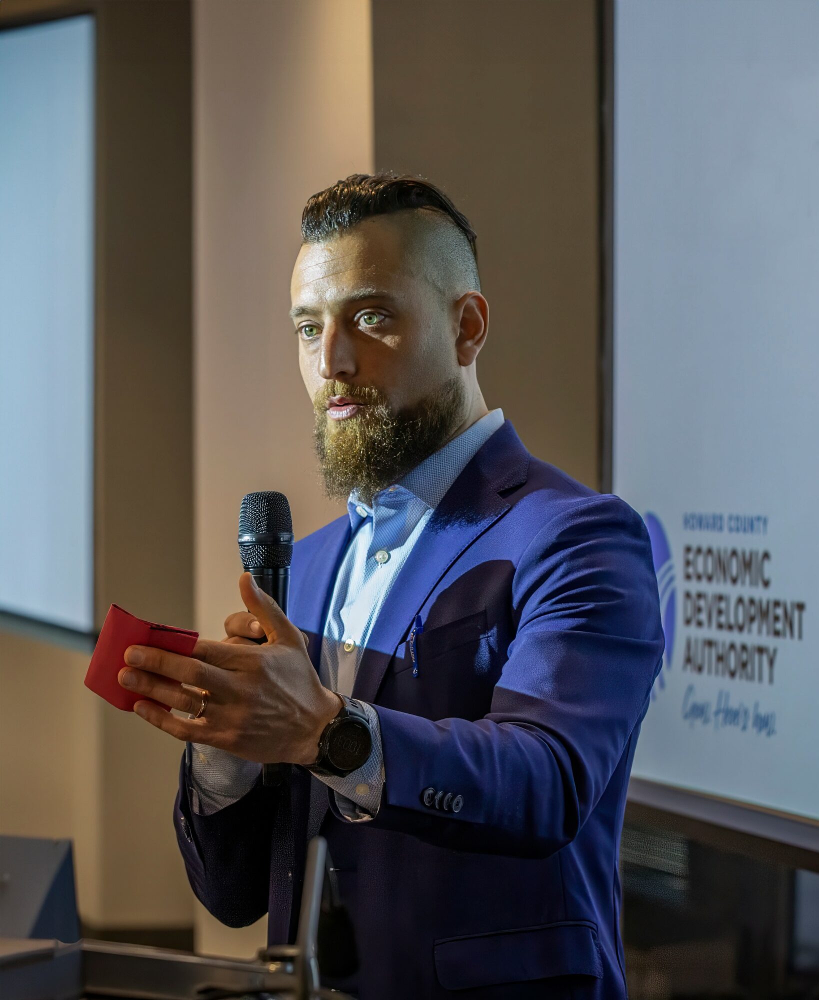 Man in blue suit holding a microphone and a red notepad, standing in front of a presentation screen.