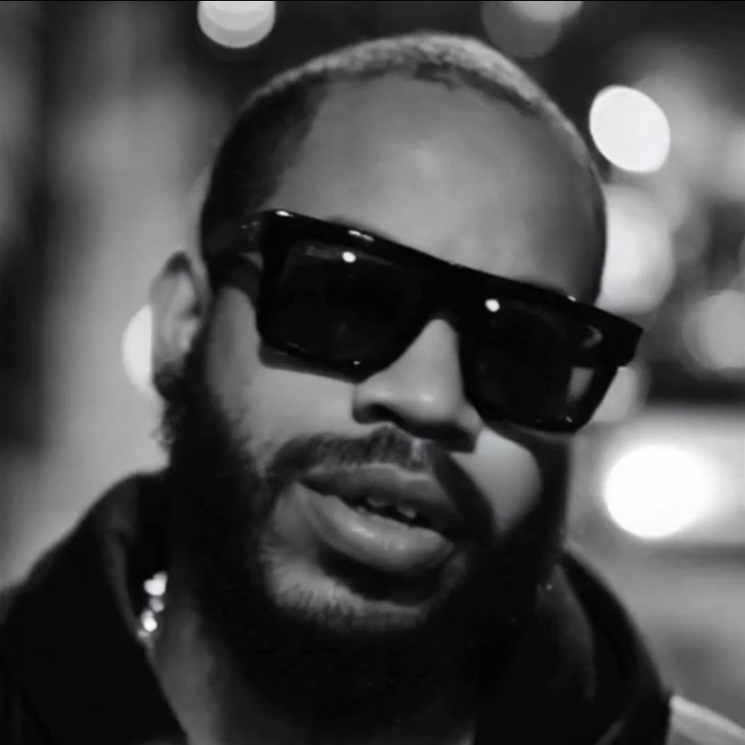 Black and white close-up of a person wearing large sunglasses and a hoodie, with blurred city lights in the background.