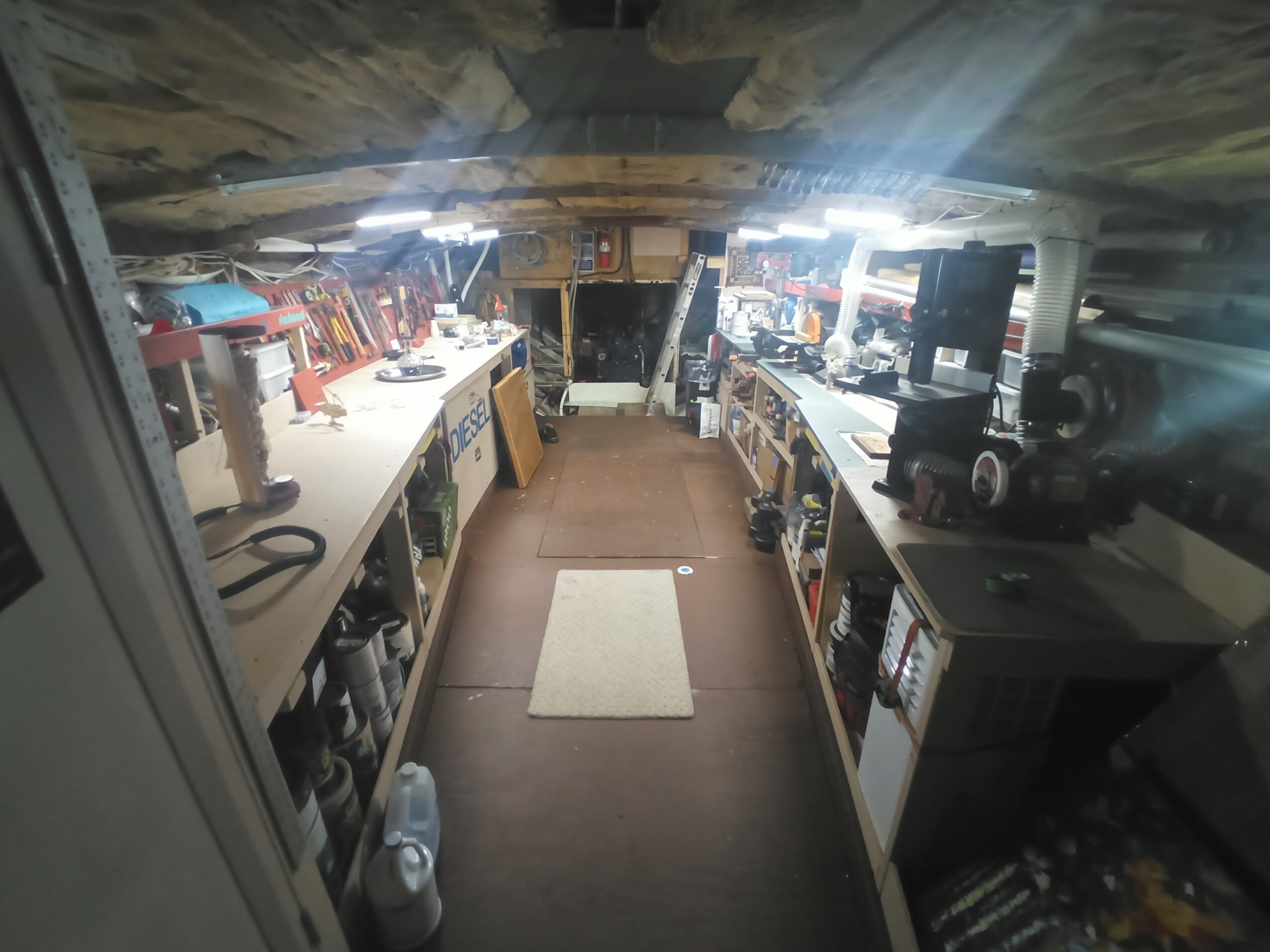 Workshop with workbenches, tools, and equipment on both sides, ceiling with exposed beams, and a small rug in the center.