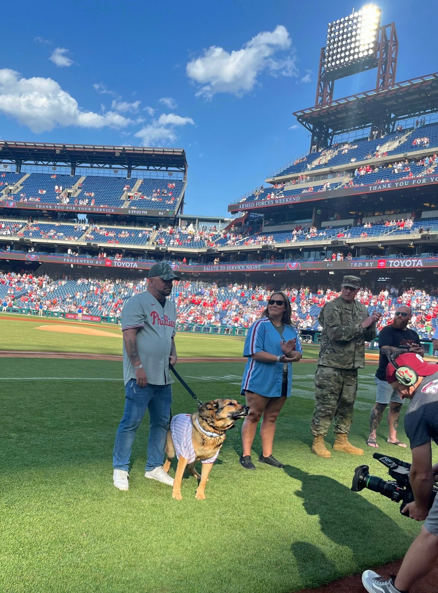 People and a dog on a baseball field with stadium seating and a bright sky in the background.