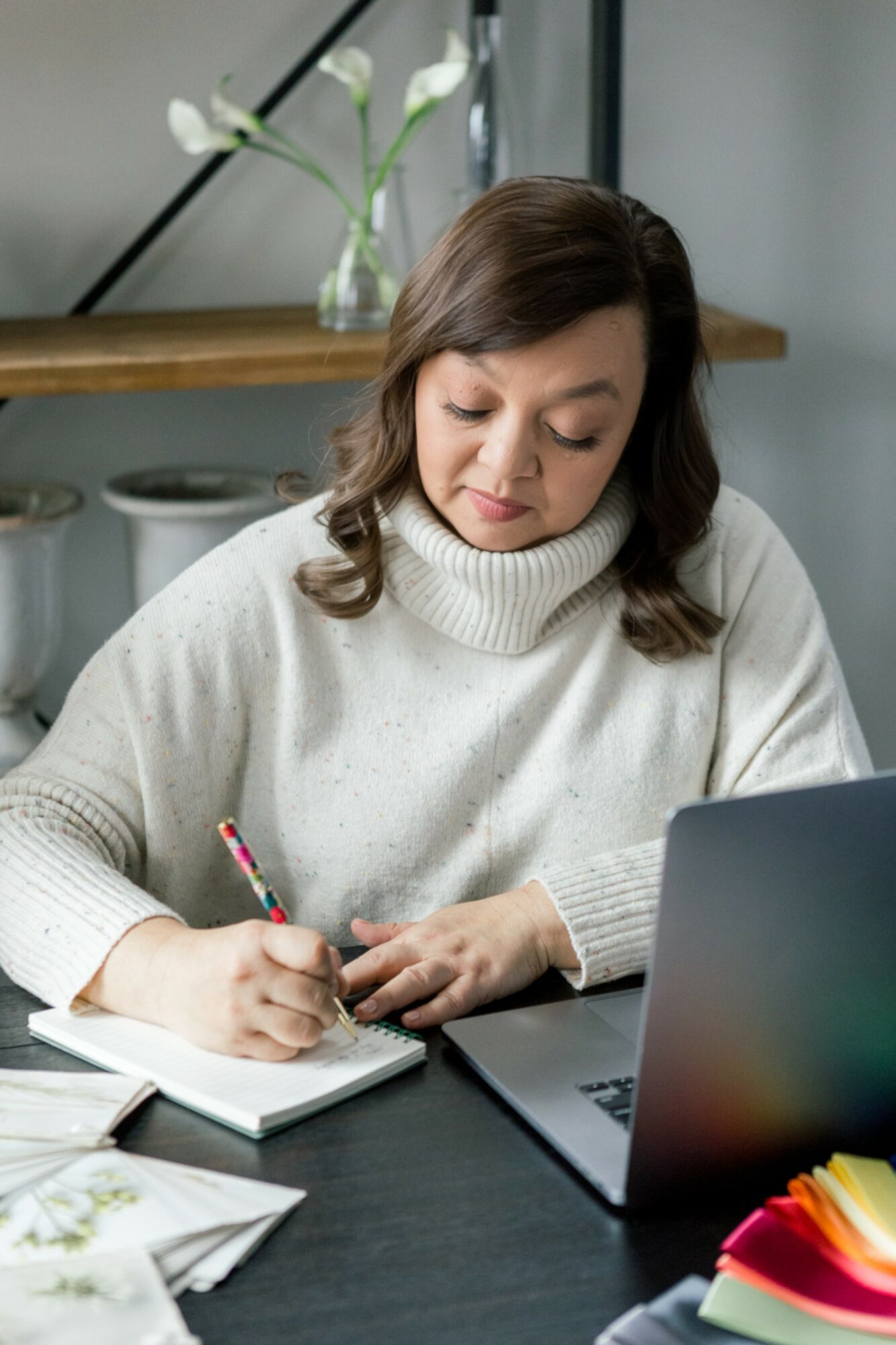 Woman in white sweater writing in notebook at desk with laptop and papers, plant in background