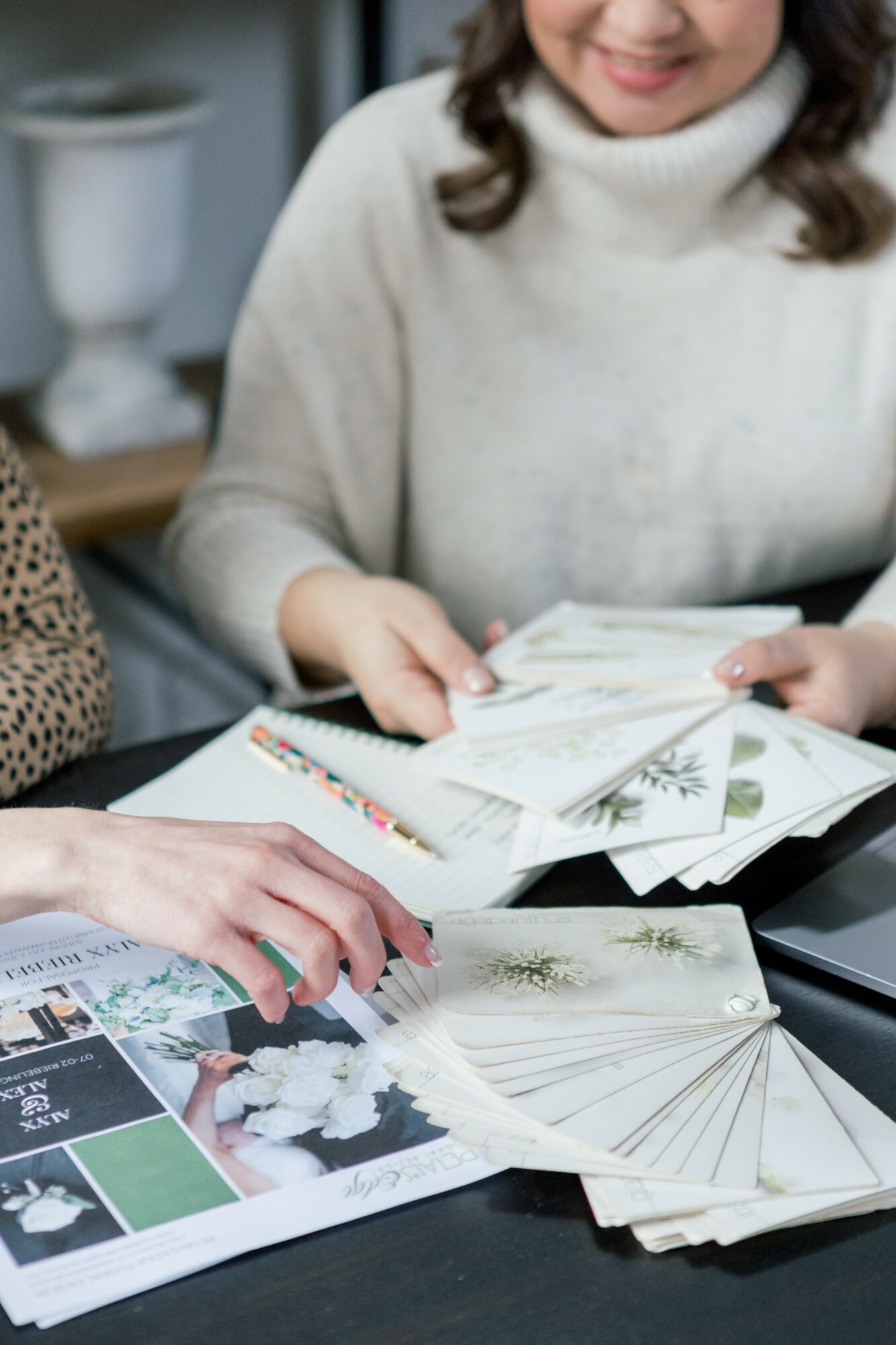 Two women look at printed materials and a fan on a table, smiling and engaging in discussion.