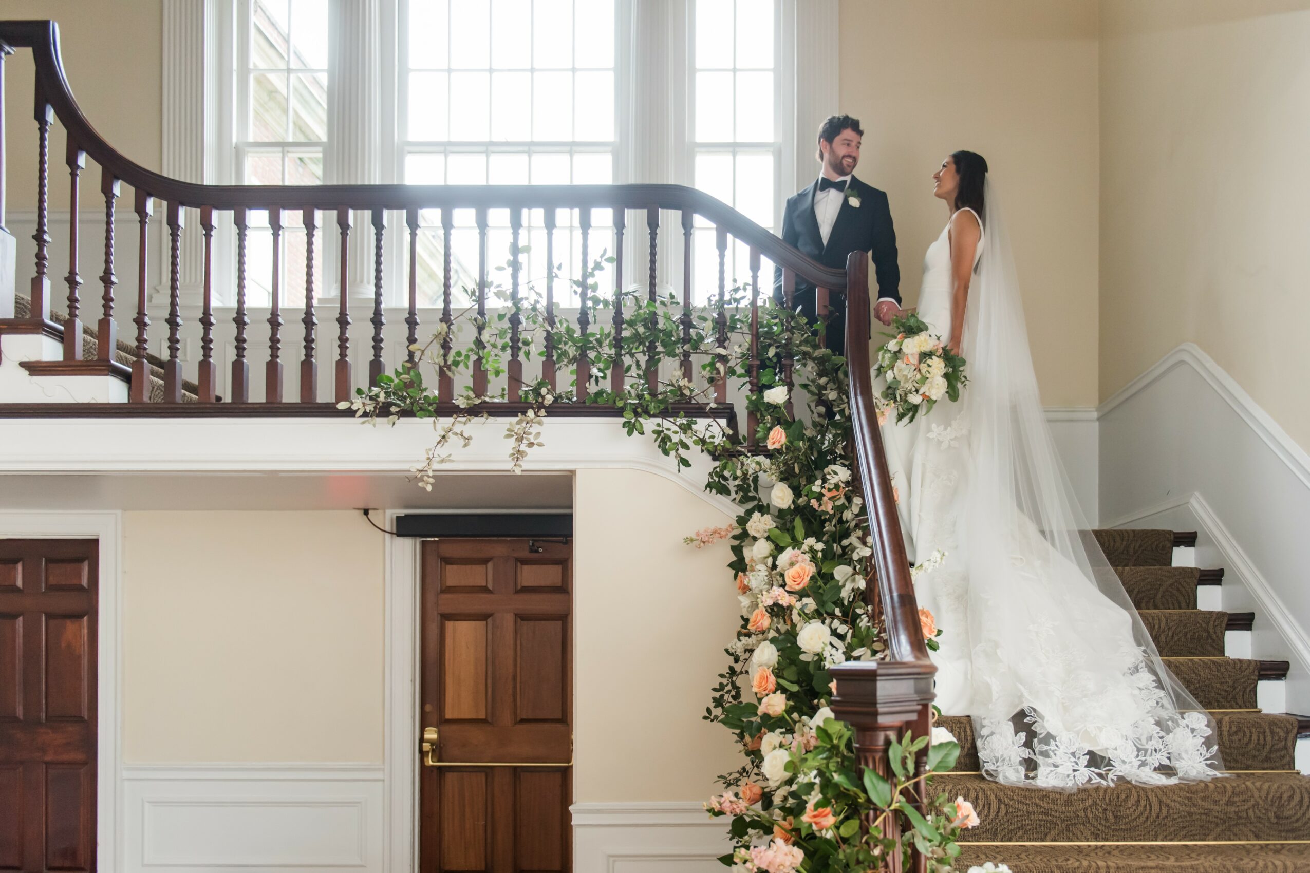 Bride and groom standing on staircase landing with floral arrangement, sunlight streaming through large windows, elegant interior decor.