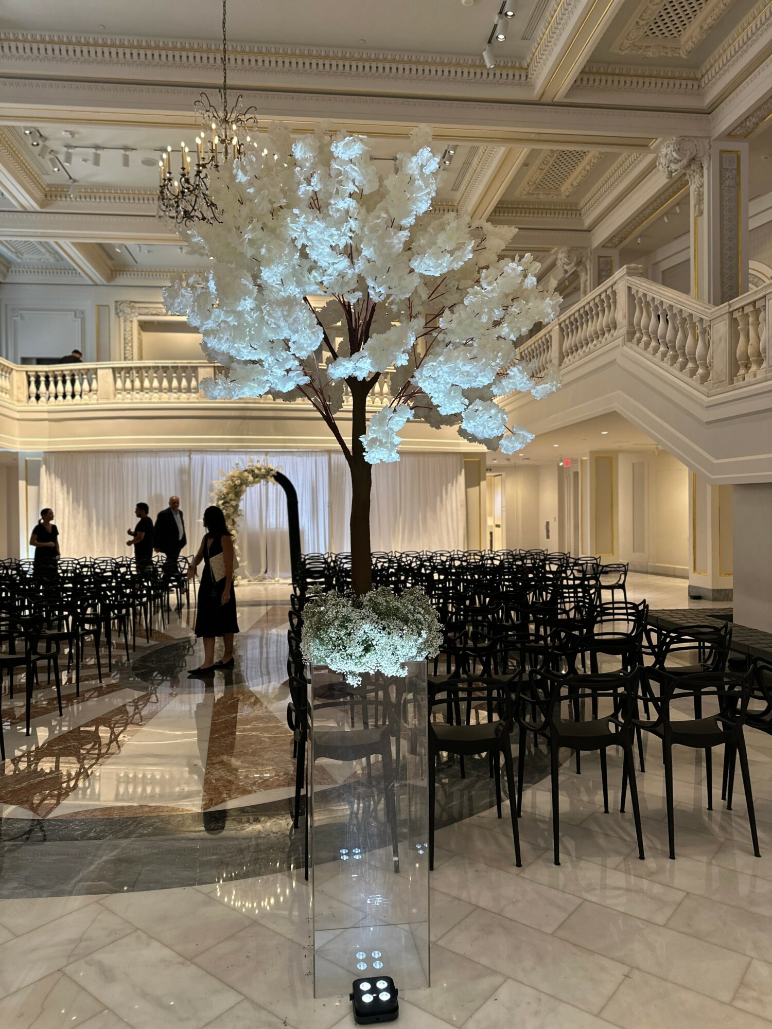 Elegant indoor event space with a large illuminated white tree, chandeliers, and rows of chairs, with a person near the tree.