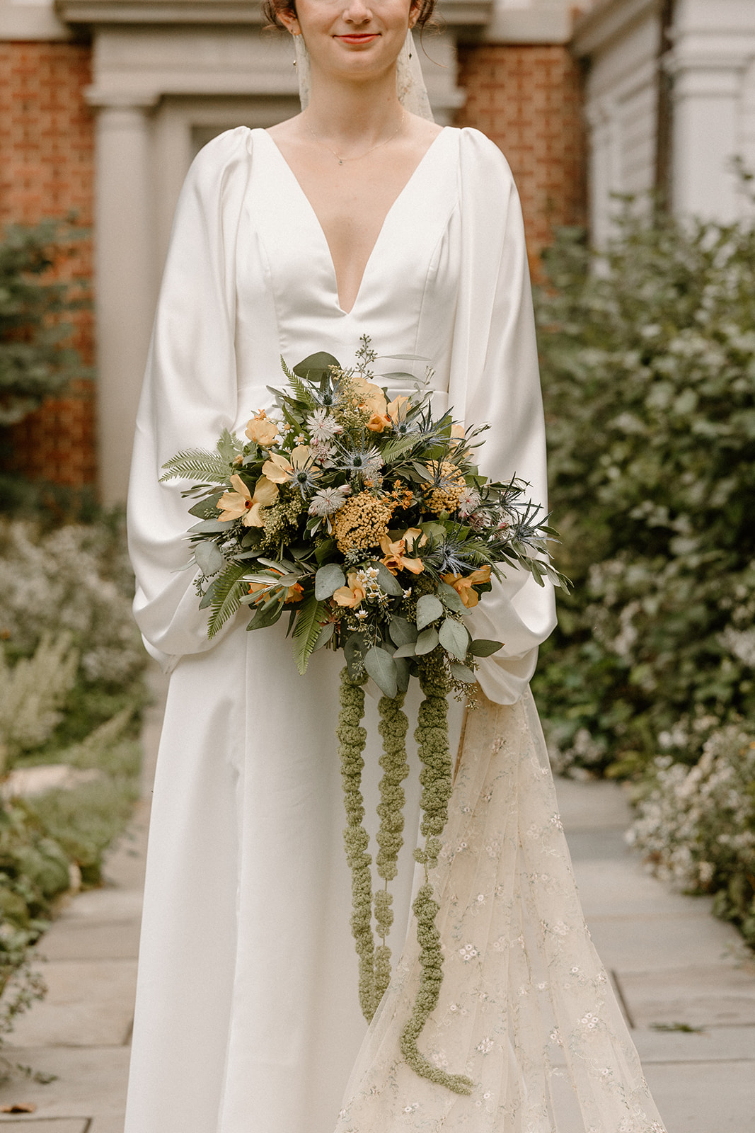 Woman in white dress holding a large bouquet of flowers outdoors, with greenery and brick building in background.