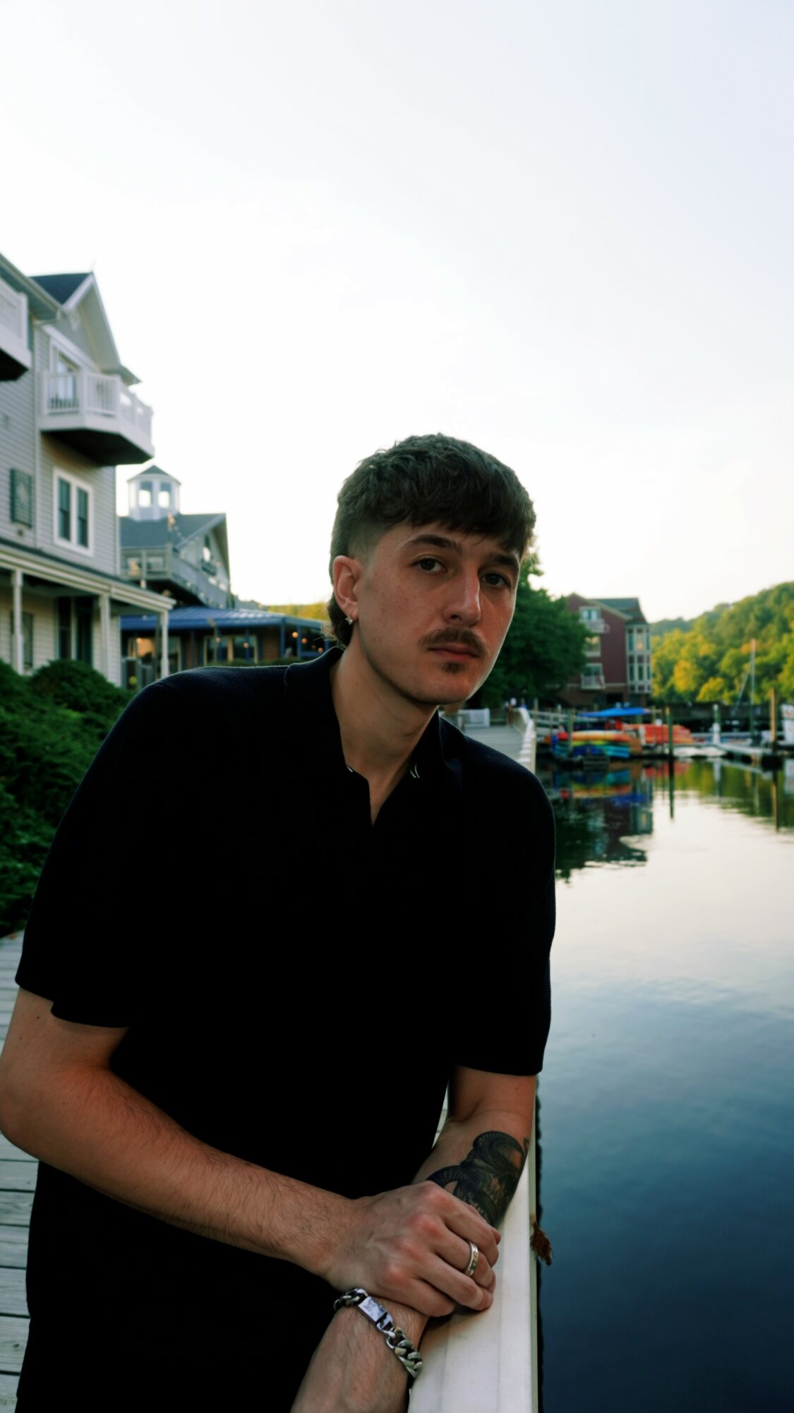 Young man with dark hair and a mustache, wearing a black shirt, standing near water with buildings in background.