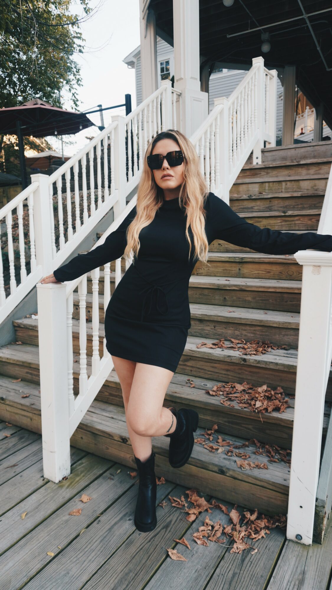 Woman in black dress and sunglasses standing on stairs outdoors, arms outstretched, with white railing and wooden steps.