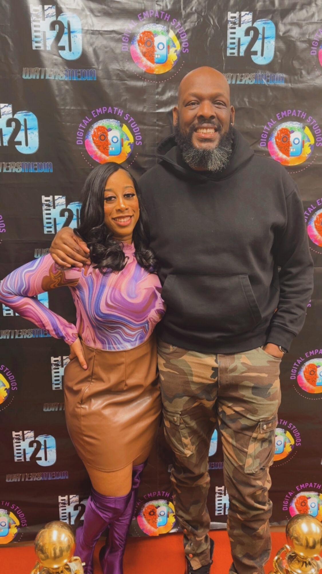 Two people standing together in front of a colorful backdrop, smiling, with one person's arm around the other.