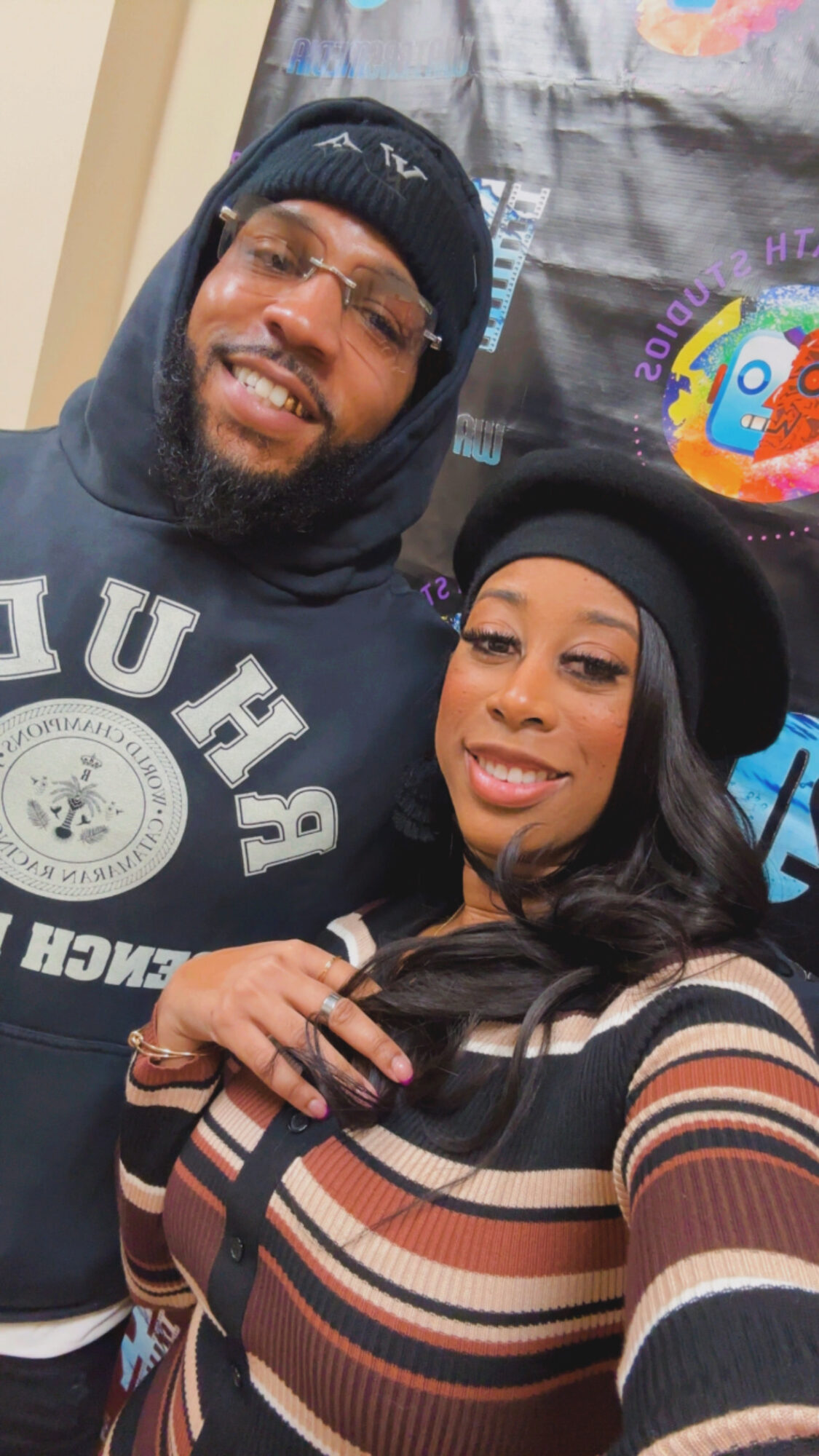 A smiling man and woman taking a selfie indoors, both wearing black hats and casual clothing.