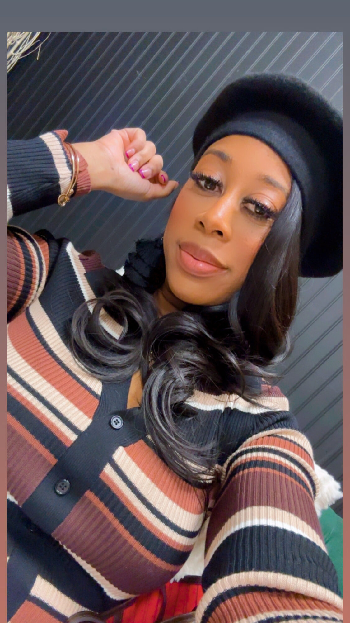 Woman wearing a striped sweater and black hat, posing with her arm raised, against a dark striped background.