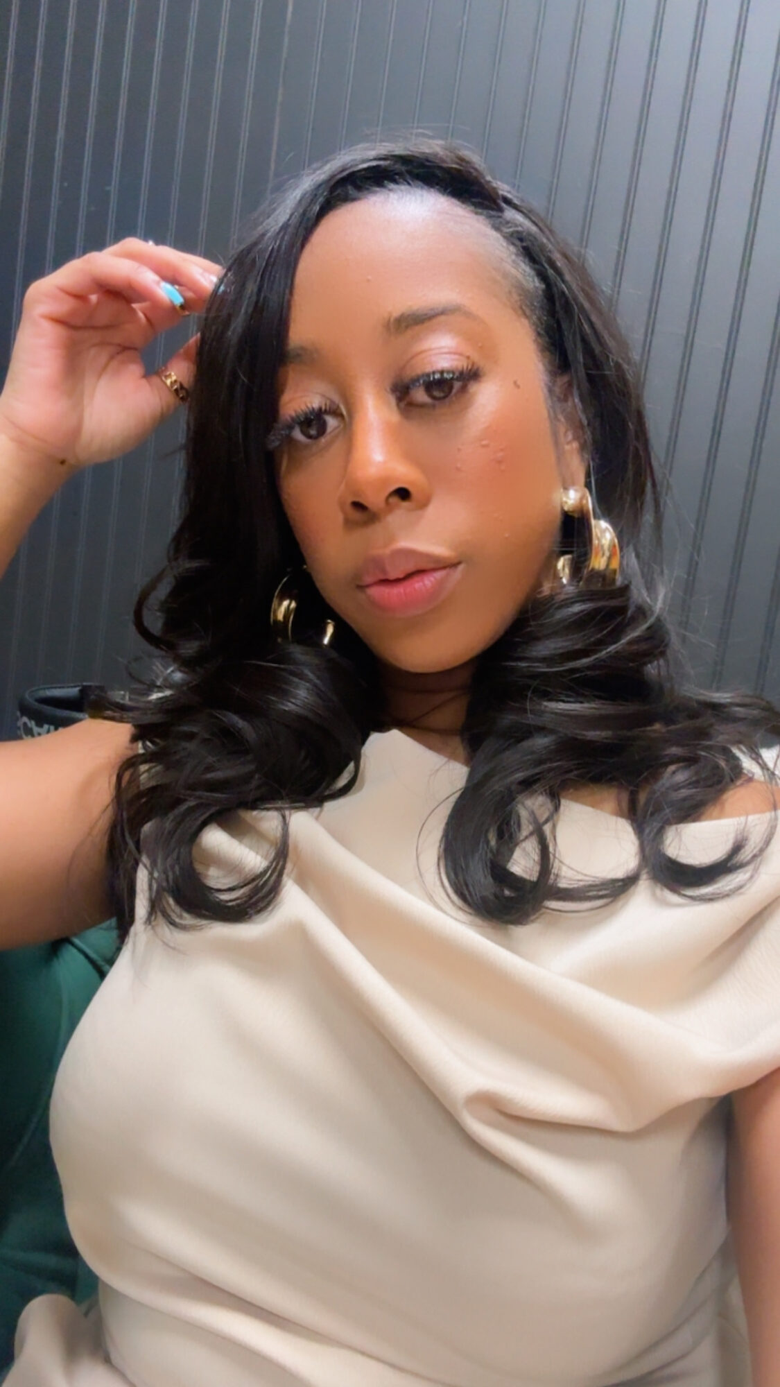Woman with dark hair and gold hoop earrings, wearing a cream top, posing indoors against a dark background.