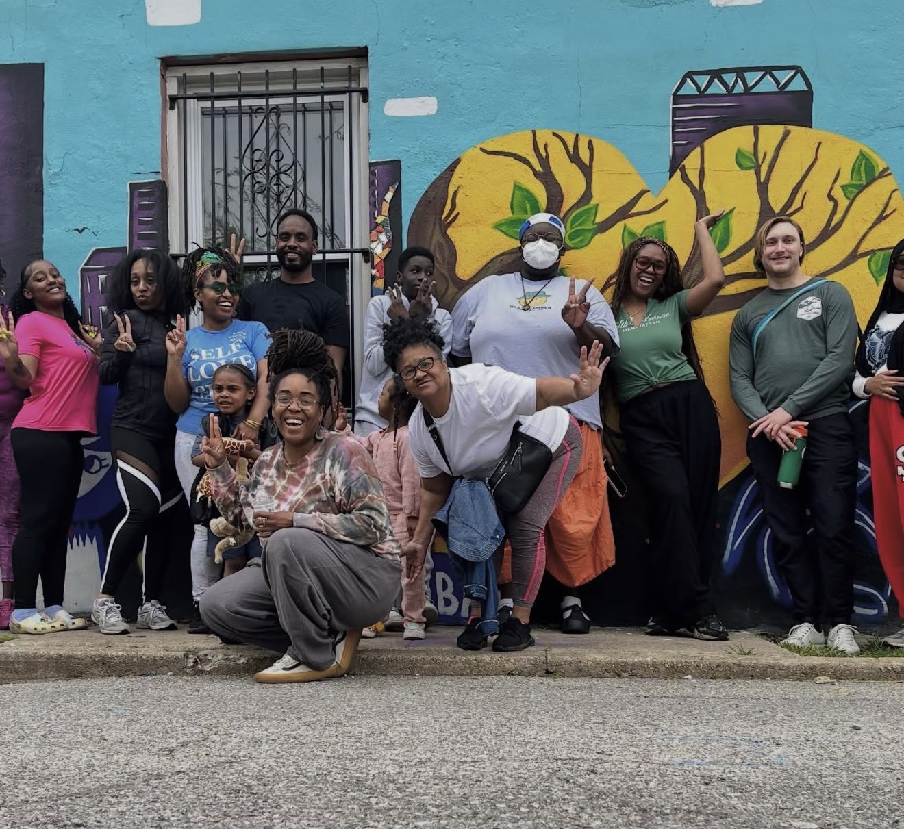 Group of people standing and kneeling in front of a colorful mural with trees and buildings, smiling and posing for the photo.