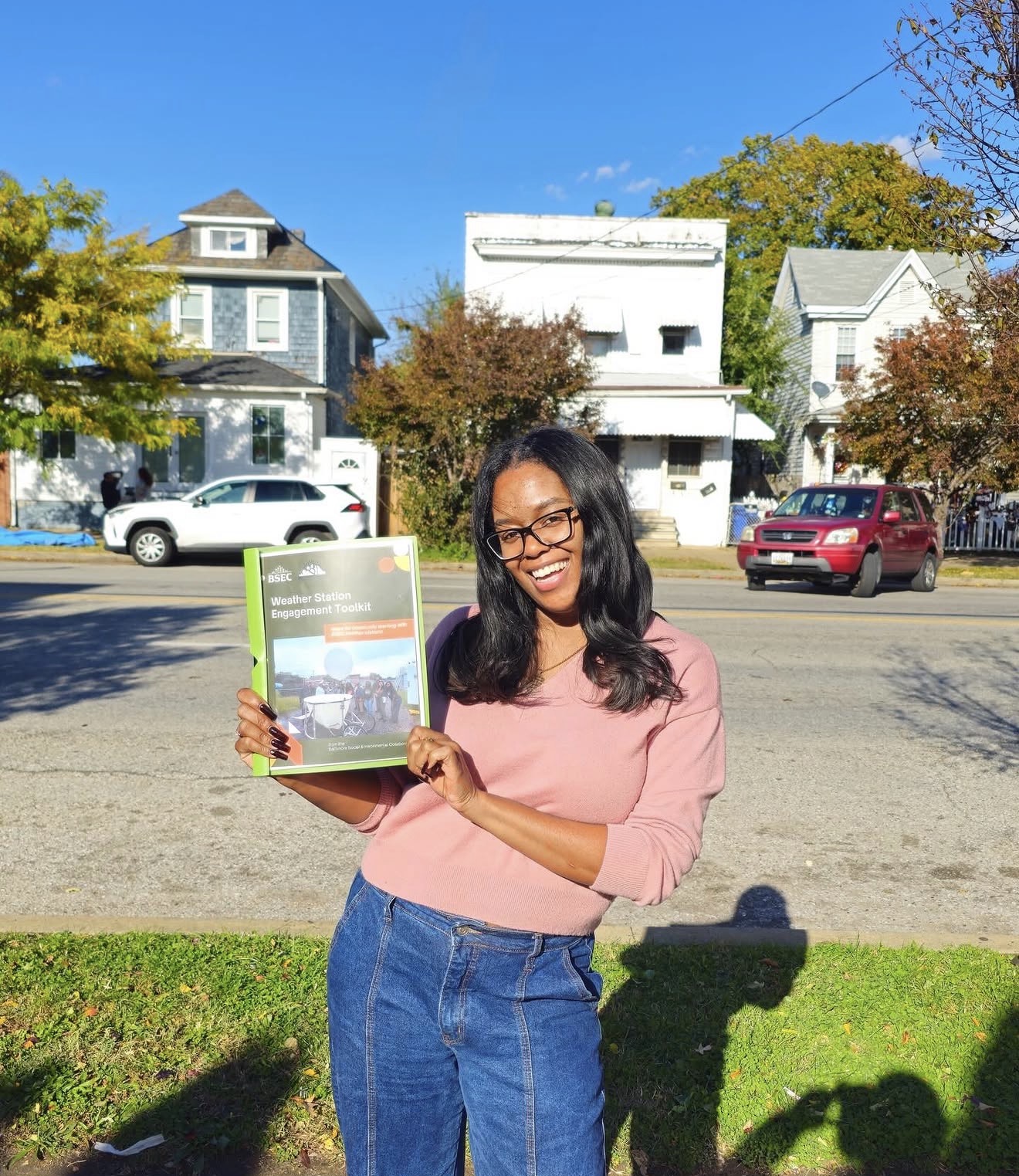 Woman with glasses holding a book outdoors on a sunny day, smiling, with houses and cars in the background.
