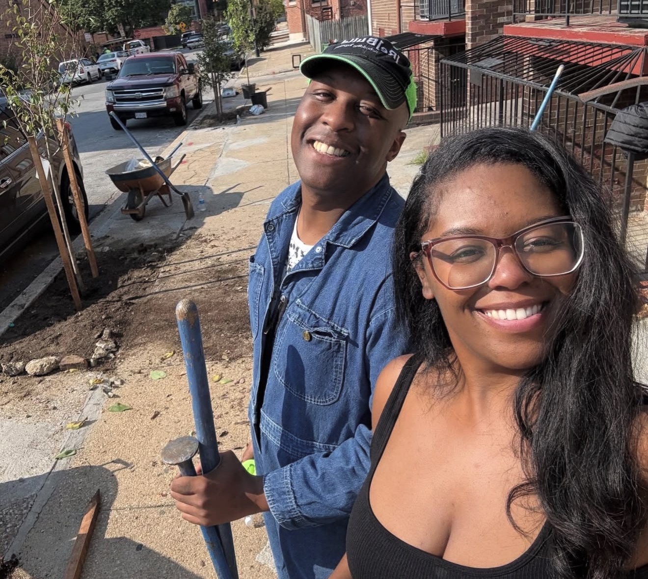 Two smiling people taking a selfie outdoors on a sunny day, with a street and buildings in the background.