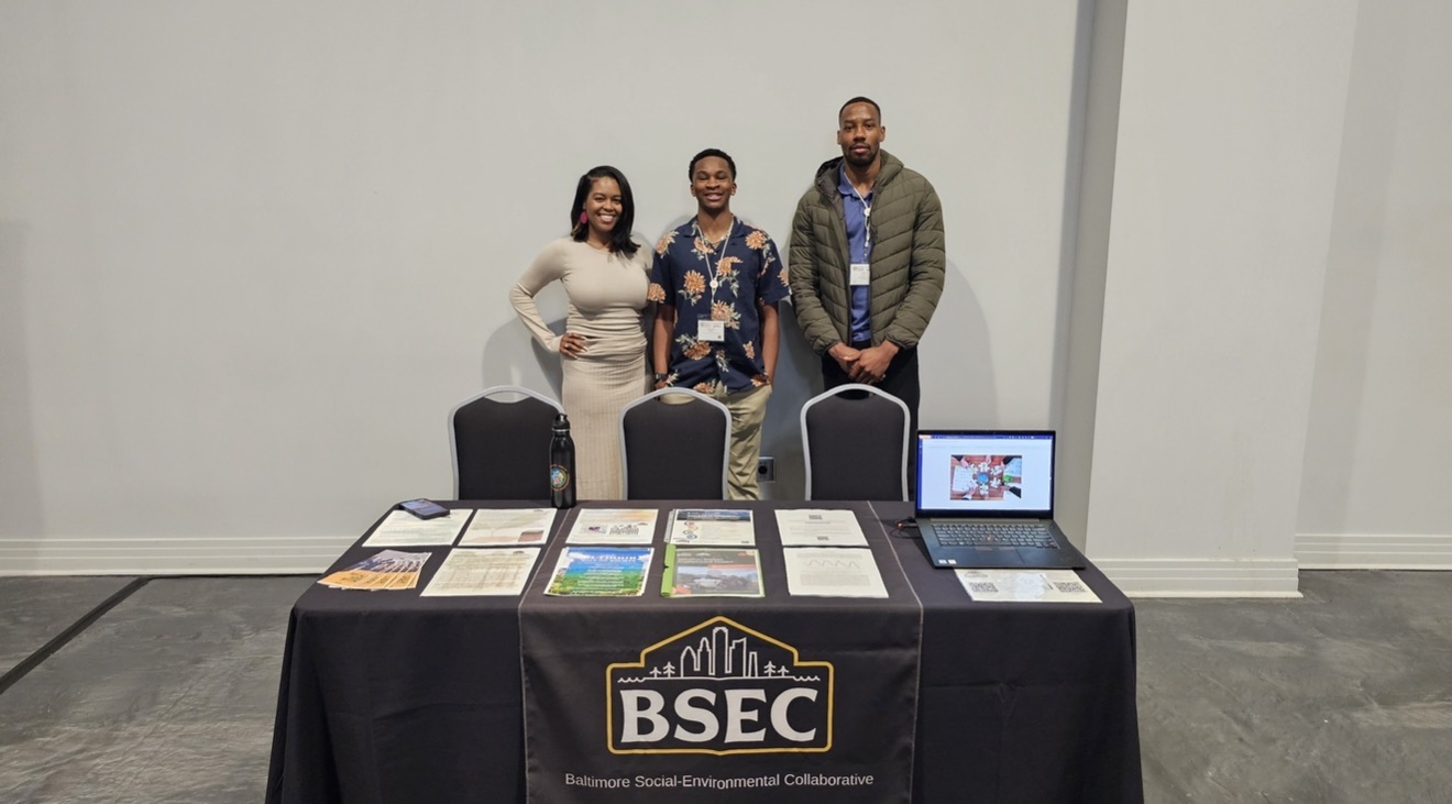 Three people standing behind a table with brochures and a laptop, in a room with white walls.