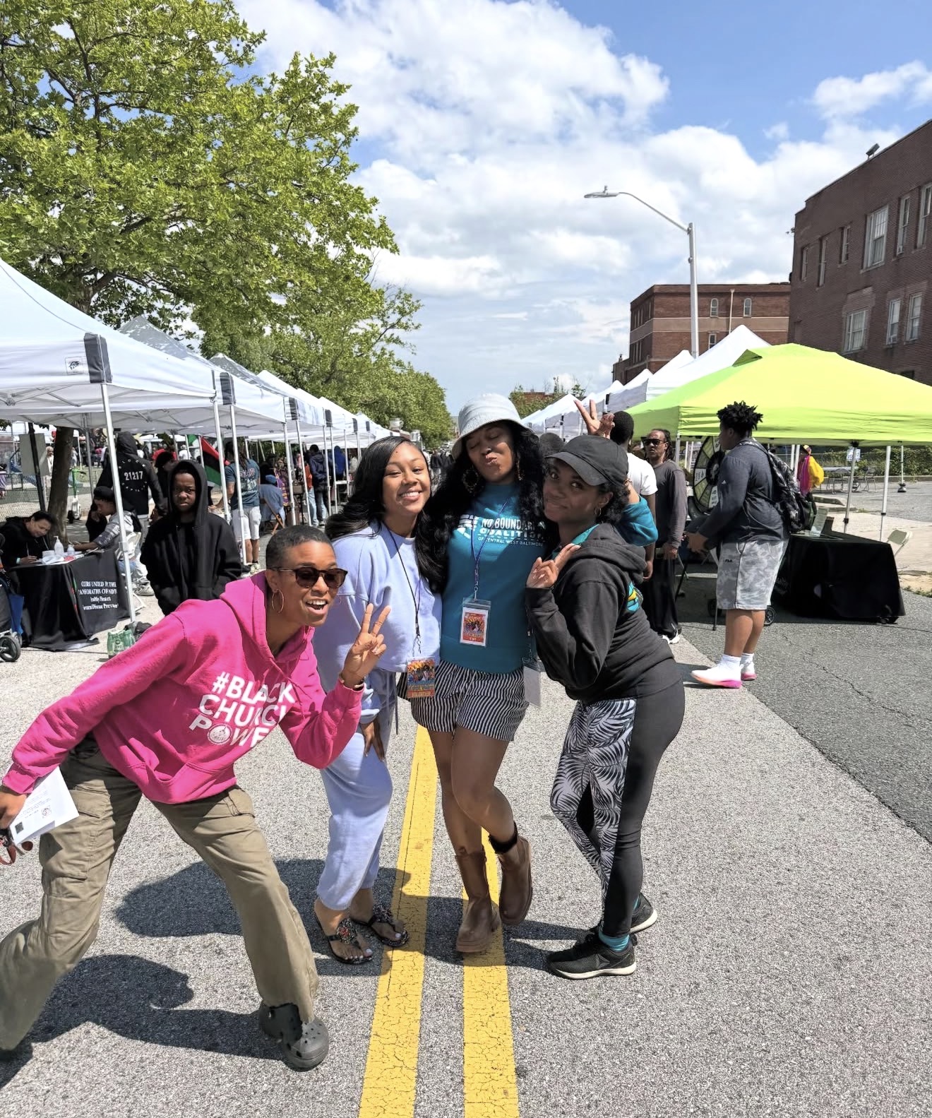 Four women standing on a street with tents and people in the background, smiling and posing for the photo.