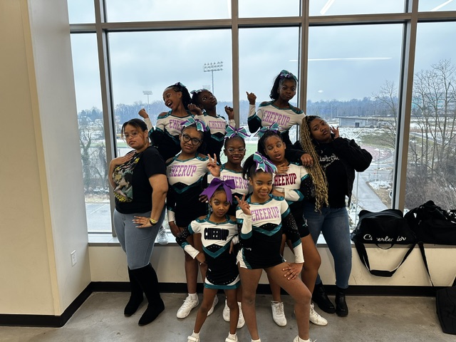 Group of children and adults posing inside near large window, some children holding signs, outdoor view visible through window