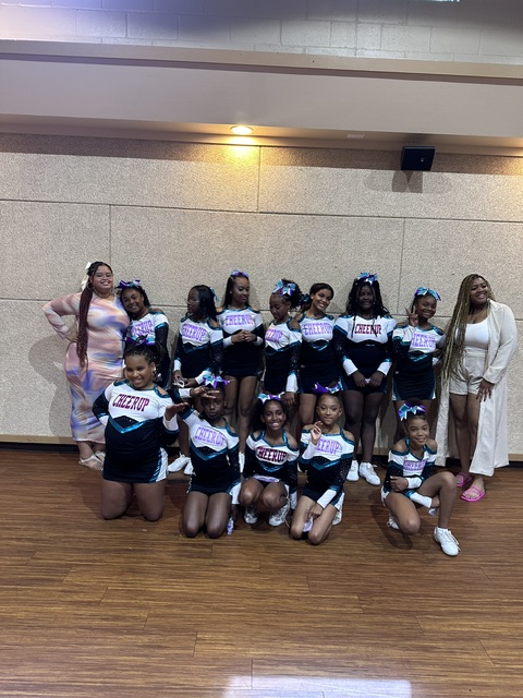 Group of young female cheerleaders in black uniforms with pink and white accents, holding pom-poms, posing indoors on a wooden floor.