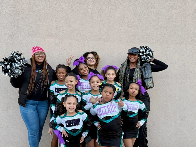 Group of young cheerleaders with two adults, smiling and holding pom-poms, standing against a plain wall.
