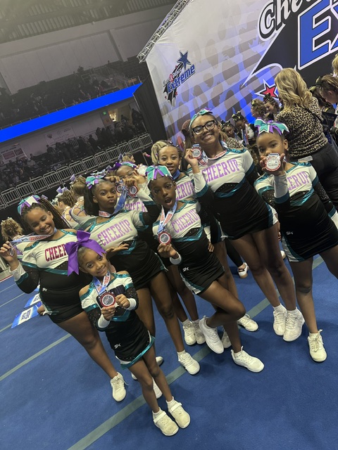 Group of young cheerleaders in colorful uniforms and pom-poms, standing on a blue floor with a banner in the background.