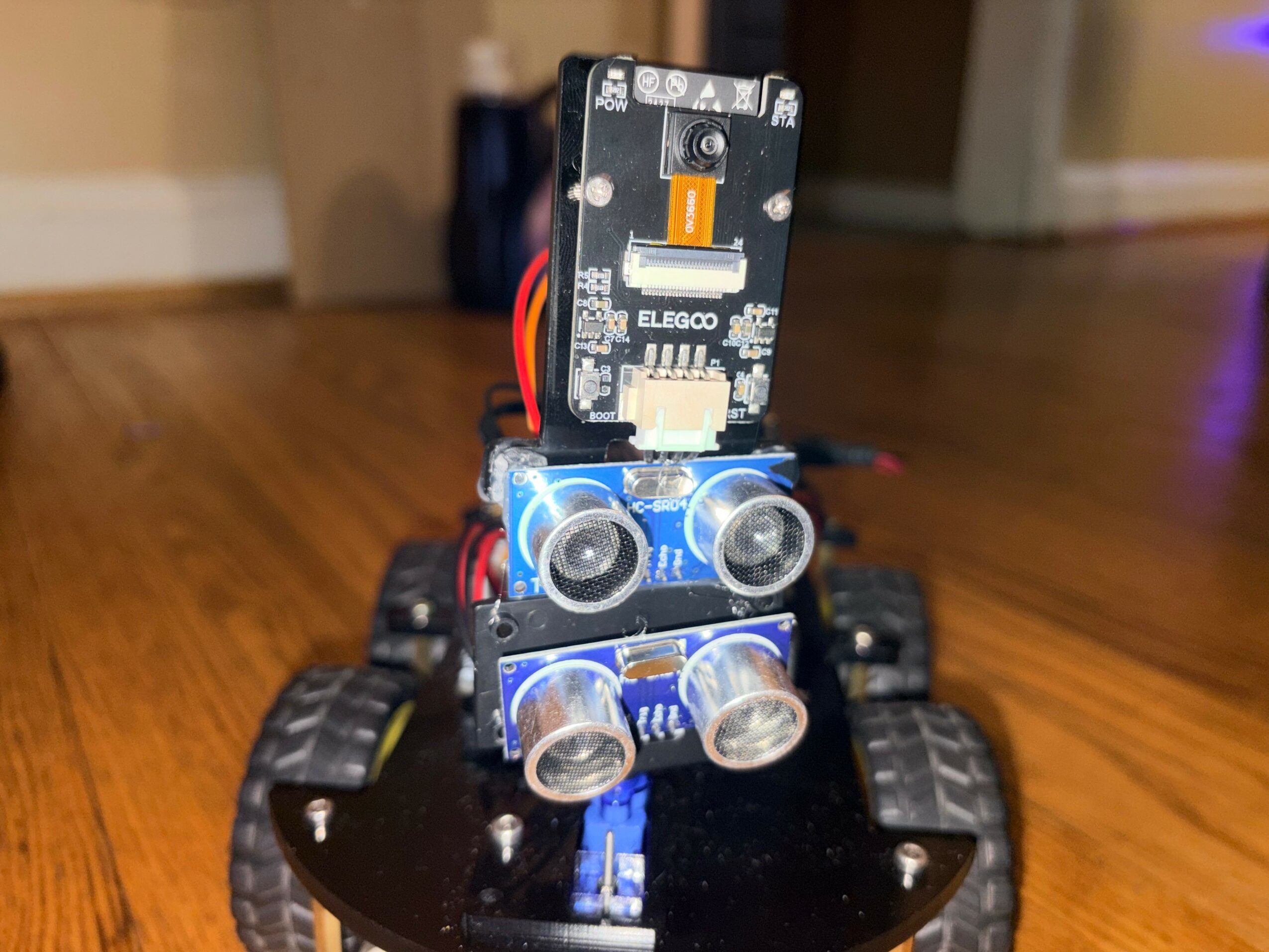Small robot with four clear plastic wheels, two illuminated blue lights, and a circuit board on top, on a wooden floor.