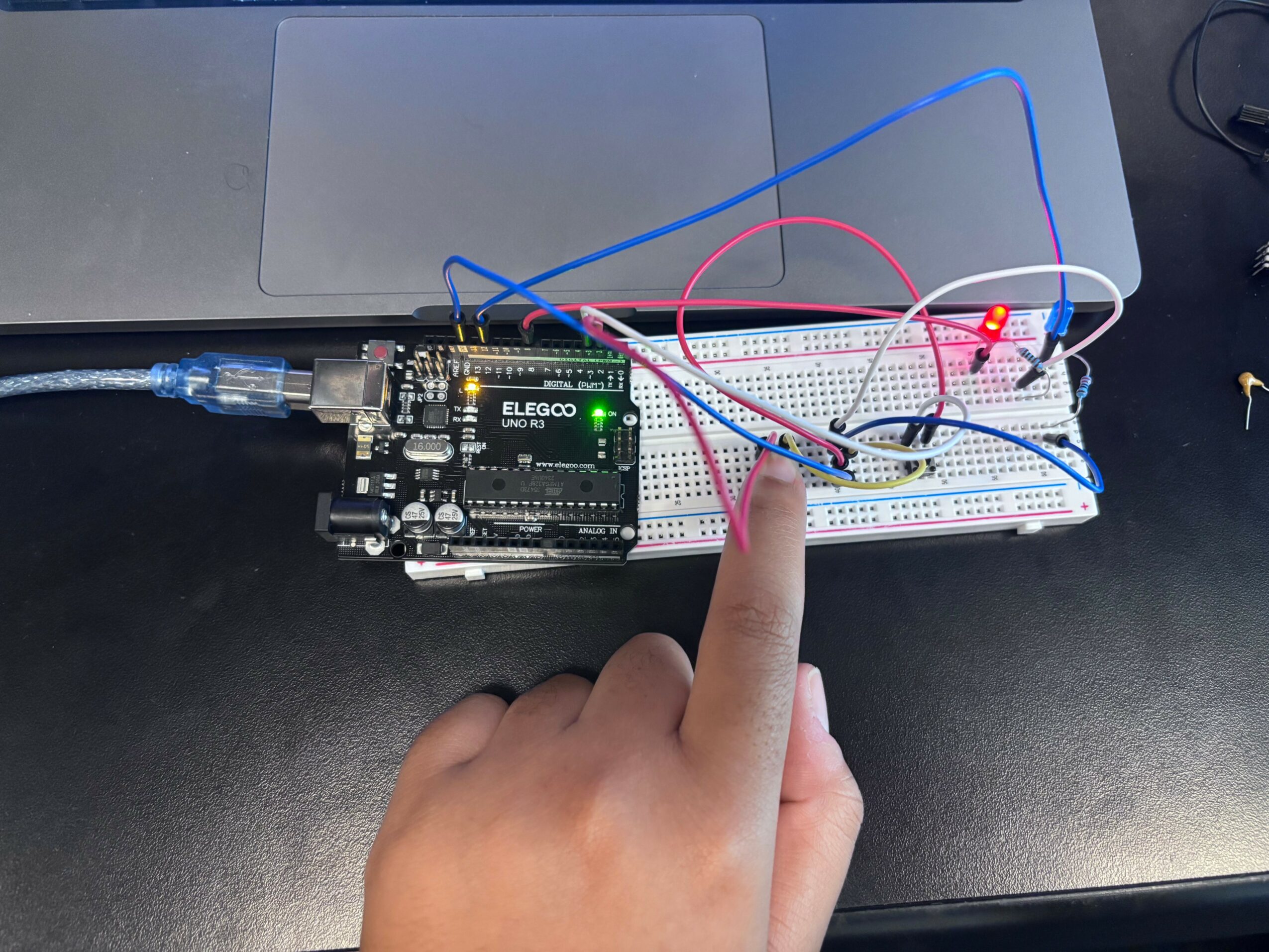 Person's finger points at a breadboard with electronic components connected to a microcontroller, on a black surface.