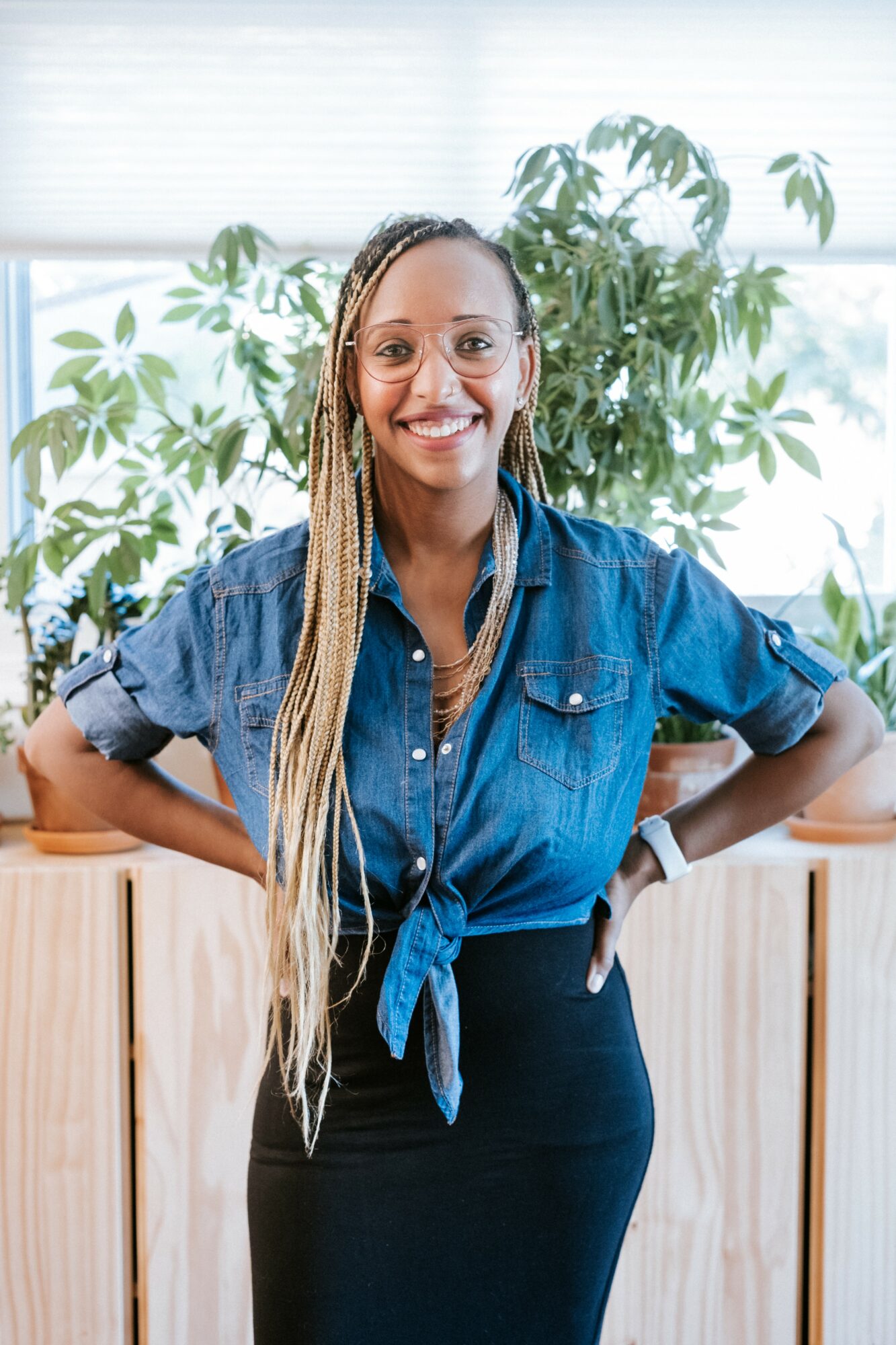 Smiling woman with long braided hair wearing a denim shirt and black skirt, standing indoors with plants in the background.