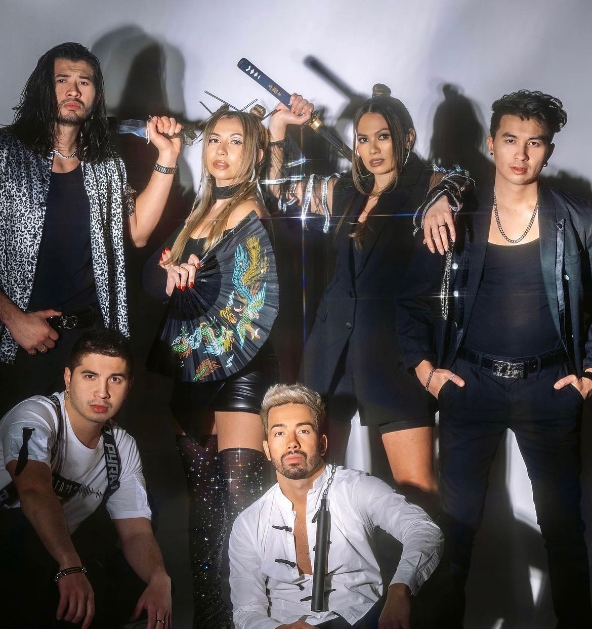 Group of six young adults posing with weapons, some with serious expressions, in a studio setting.