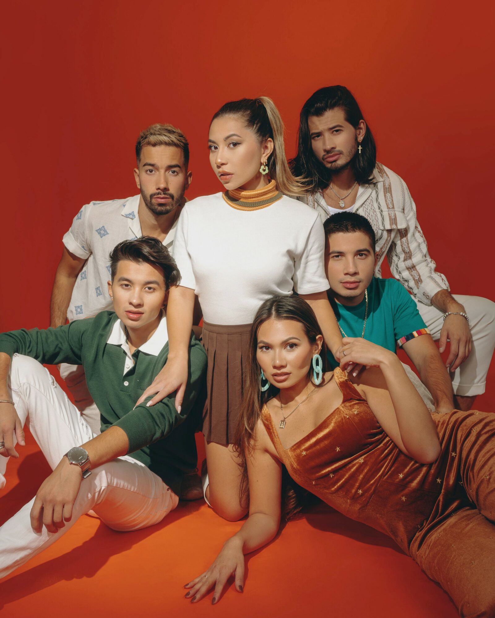 Group of six people posing against a red background, some sitting and some standing, dressed in casual and stylish clothing.