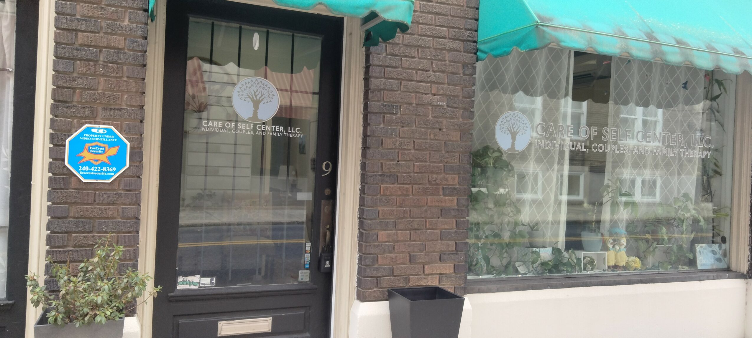 Brick building with a door and large window, green awnings, and potted plants outside.