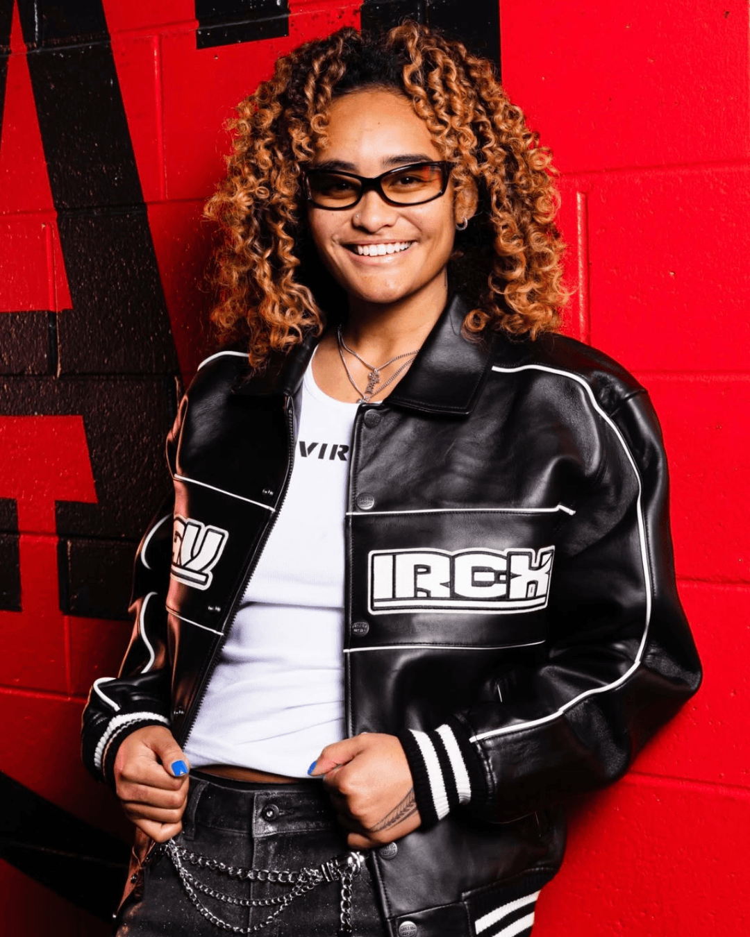 Smiling woman with curly hair and glasses, wearing a black leather jacket and white shirt, standing against a red wall.