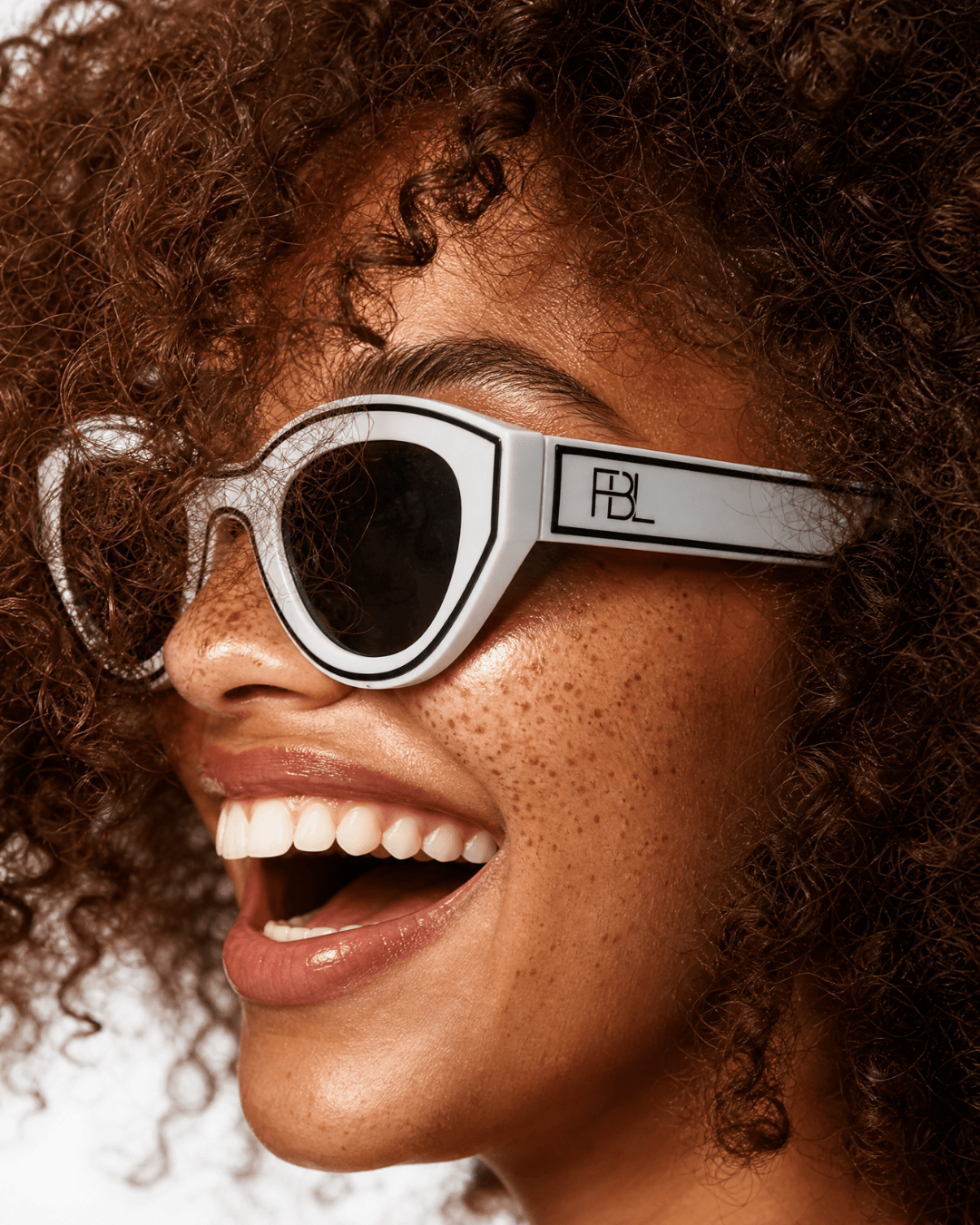 Close-up of a smiling person with curly hair wearing sunglasses, showing teeth and freckles.