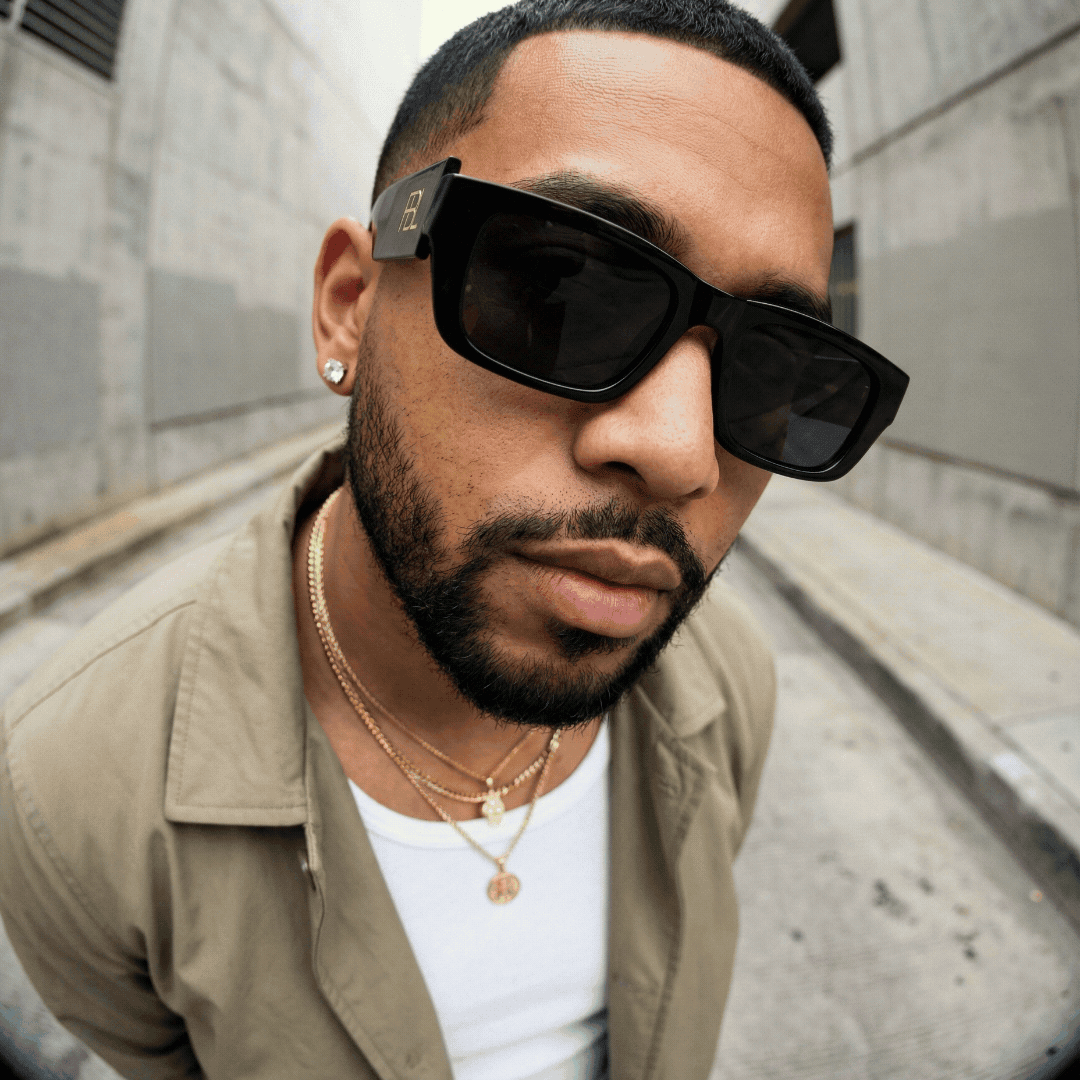 Close-up of a man wearing large sunglasses, earrings, layered necklaces, and a beige jacket in an urban setting.