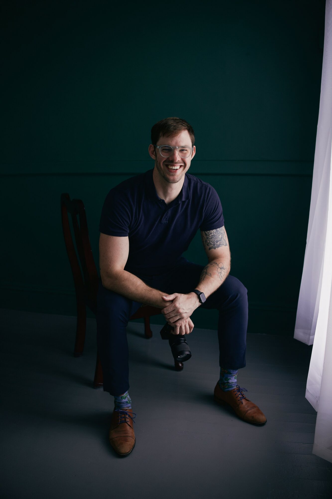 Man sitting on a chair, smiling, in a dark room with a green wall and white curtain, hands clasped, casual clothing.