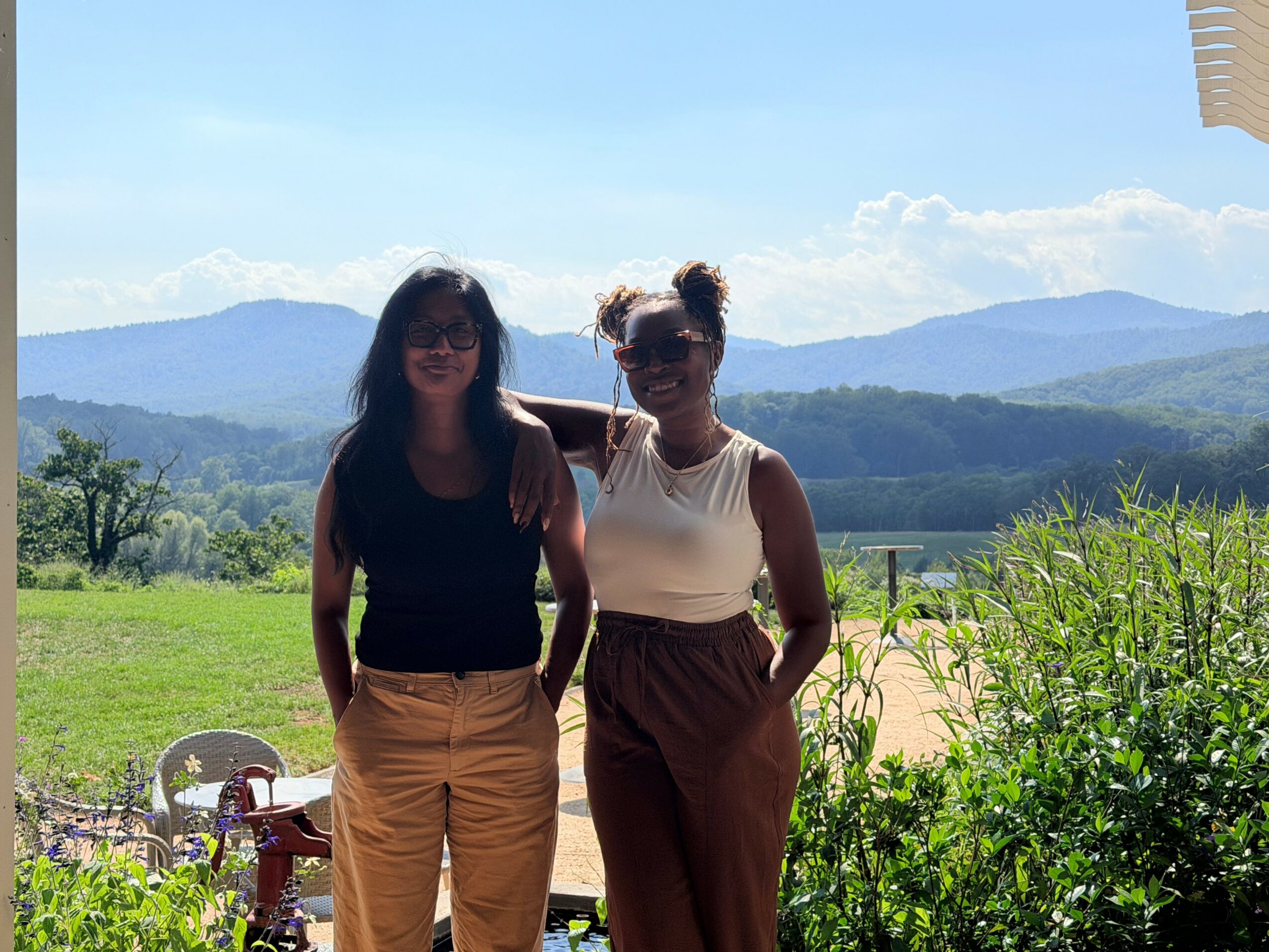 Two women stand outdoors with mountains and blue sky in background, one wearing sunglasses and the other with a bun hairstyle.