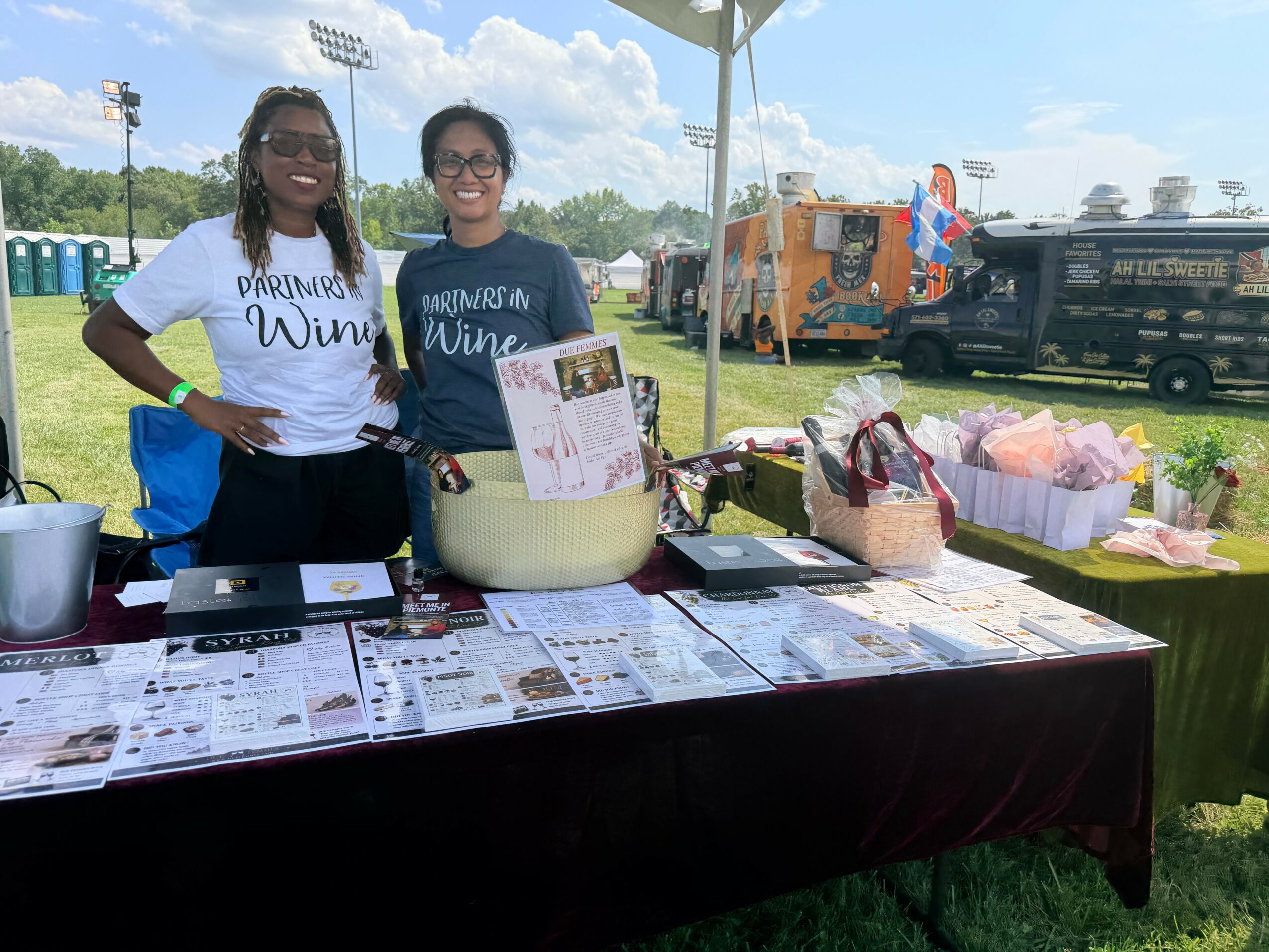 Two women stand behind a table with flyers and a large container, outdoors under a tent, with trucks and a field in background.