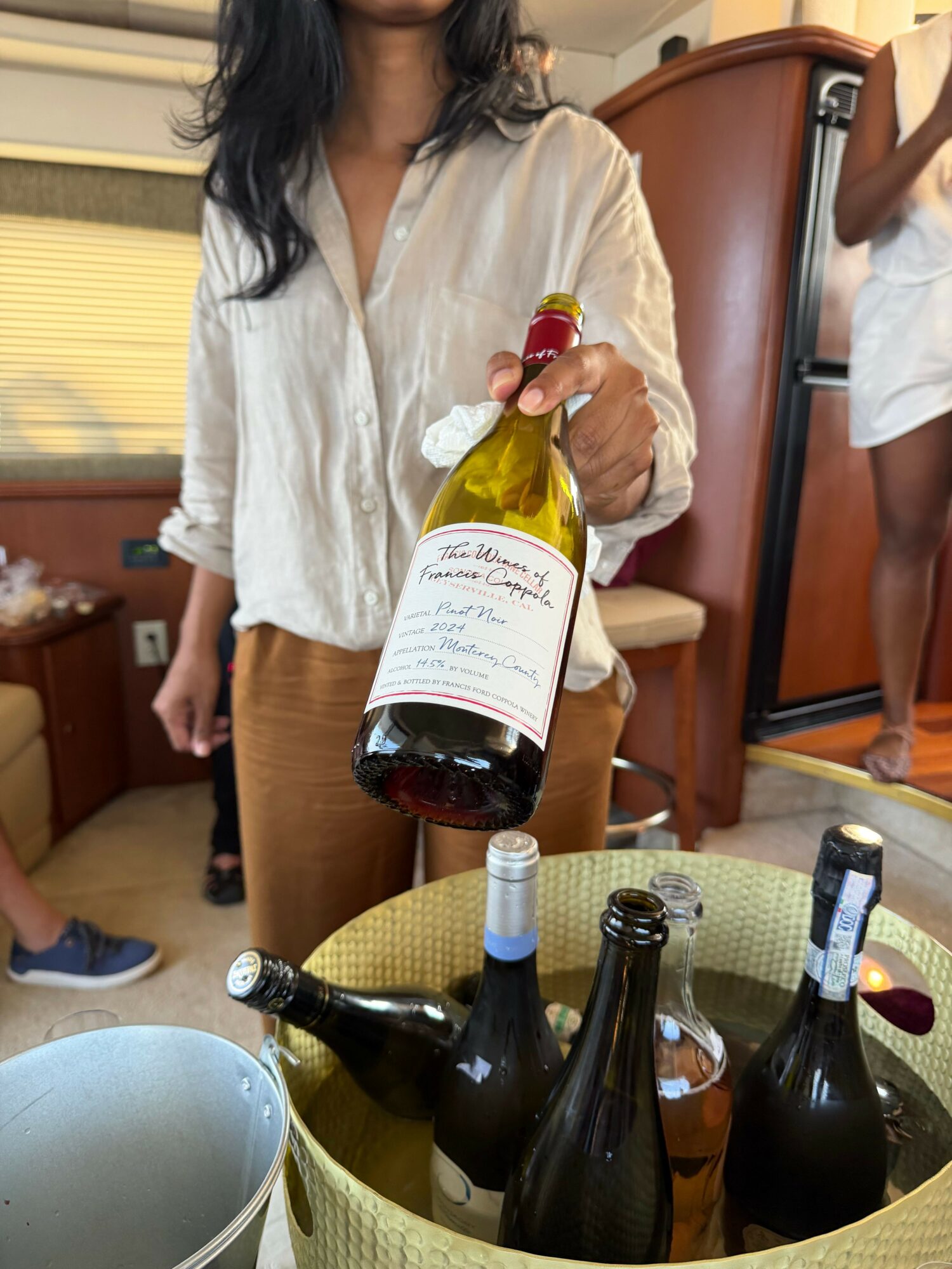 Person pouring wine from a bottle into glasses on a table, with several wine bottles nearby.