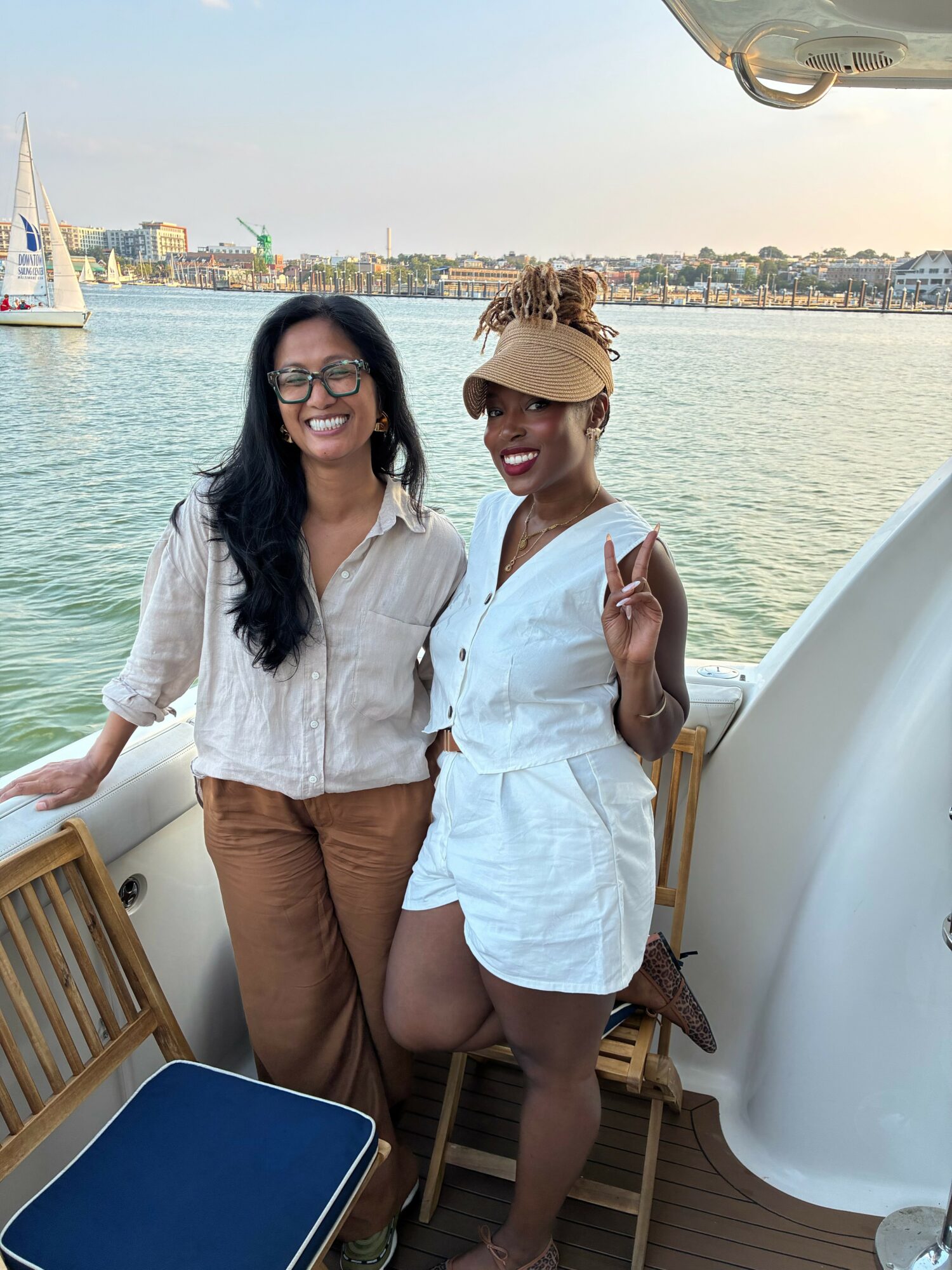 Two women smiling on a boat with water and cityscape in background, one making a peace sign, sunny day.