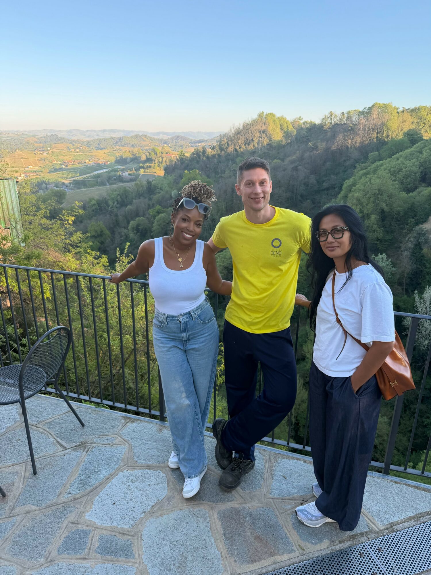 Three people standing outdoors on a balcony with a scenic landscape of trees and hills in the background.