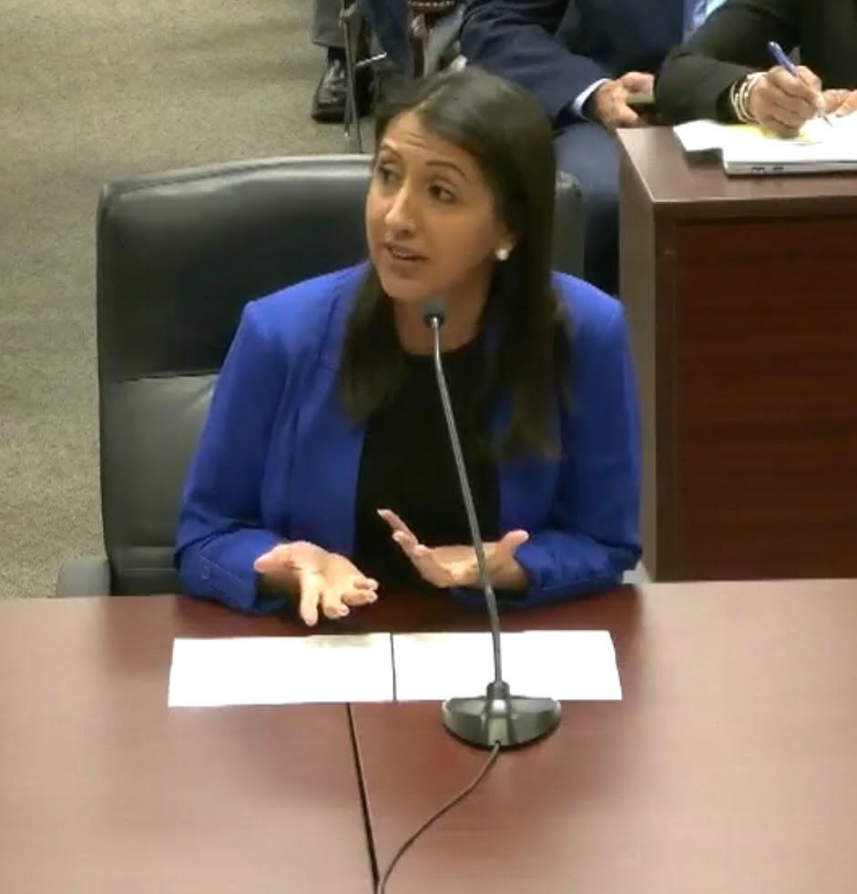 Woman in blue blazer speaking into a microphone at a desk, with people in the background taking notes.