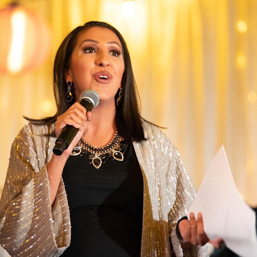 Woman speaking into a microphone, holding papers, wearing a sparkly jacket, with a warm yellow background.