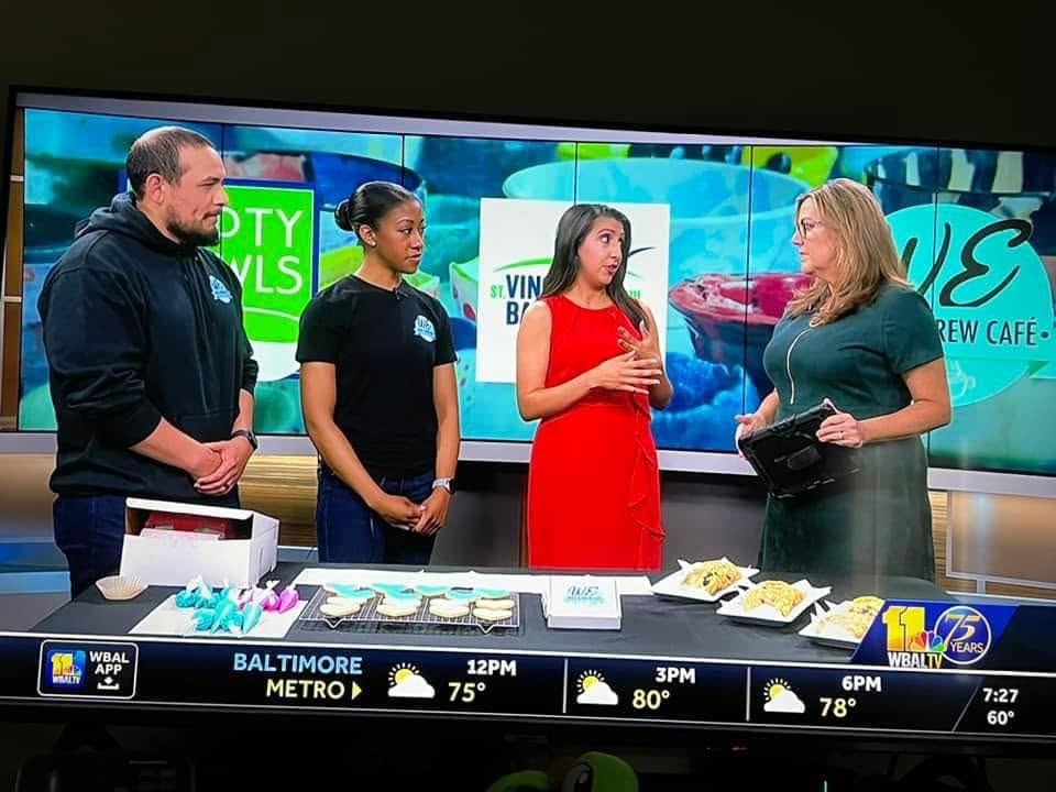 Four women stand in a TV studio with a large screen behind them displaying graphics and text. They are engaged in conversation.