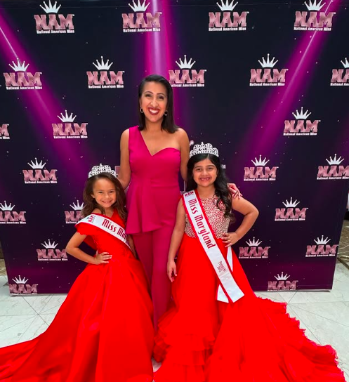 Woman and two young girls wearing crowns and sashes, standing in front of a purple backdrop with logos, smiling.